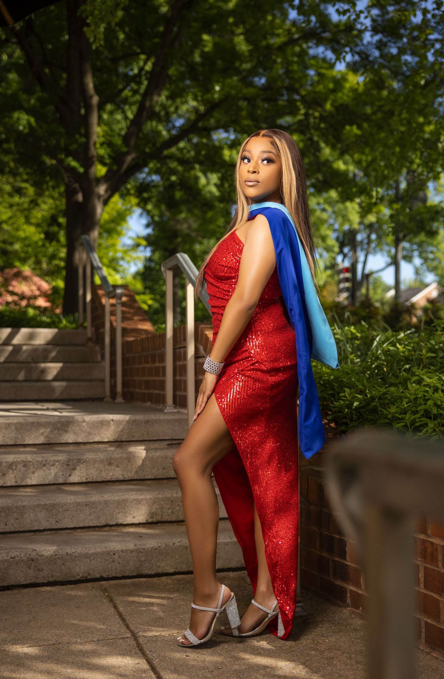 A person in a sparkly red dress and blue backpack poses on outdoor stone steps in dappled sunlight.