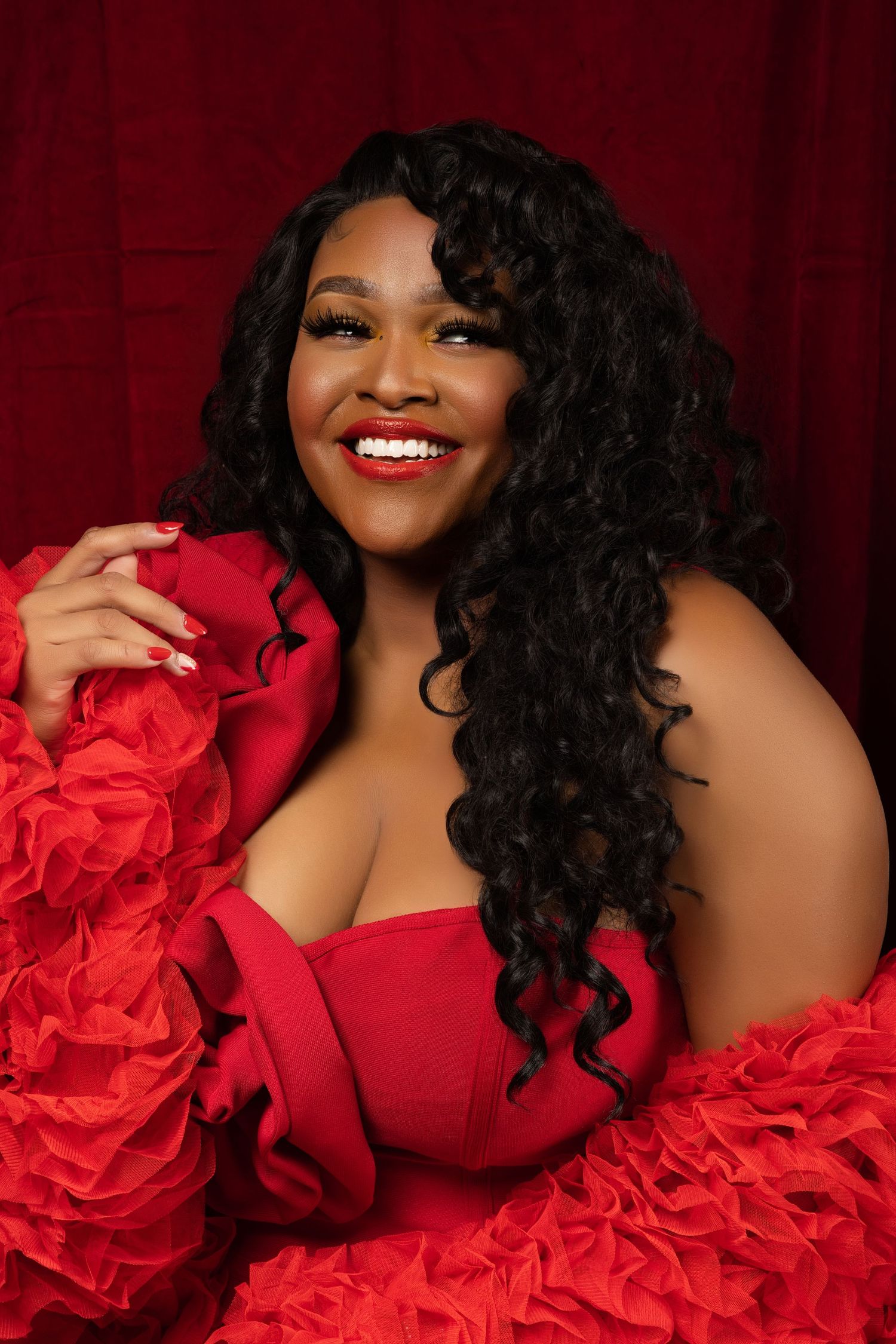 Glamorous portrait featuring red ruffled dress and curly hair against burgundy backdrop.