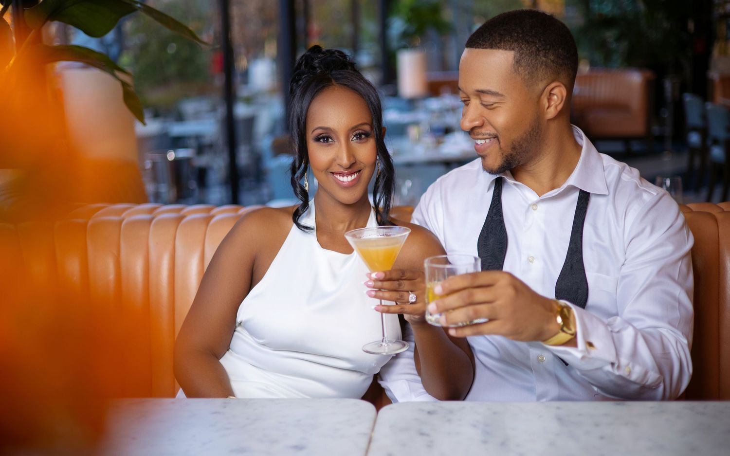 A couple in white and business attire sit at a table sharing drinks and laughing together.