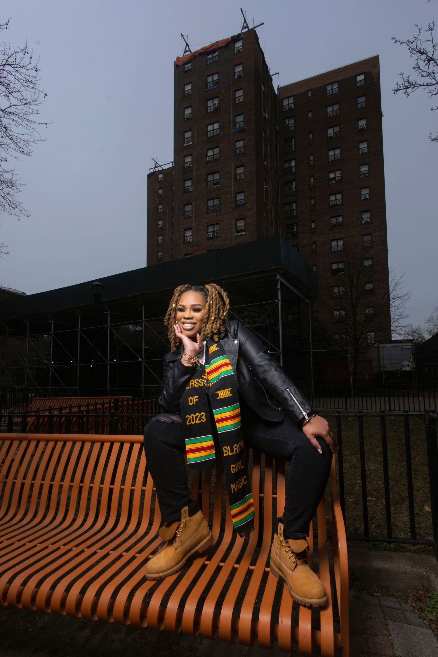 A person in a black jacket and colorful scarf sits on a wooden bench outside a tall brown apartment building.