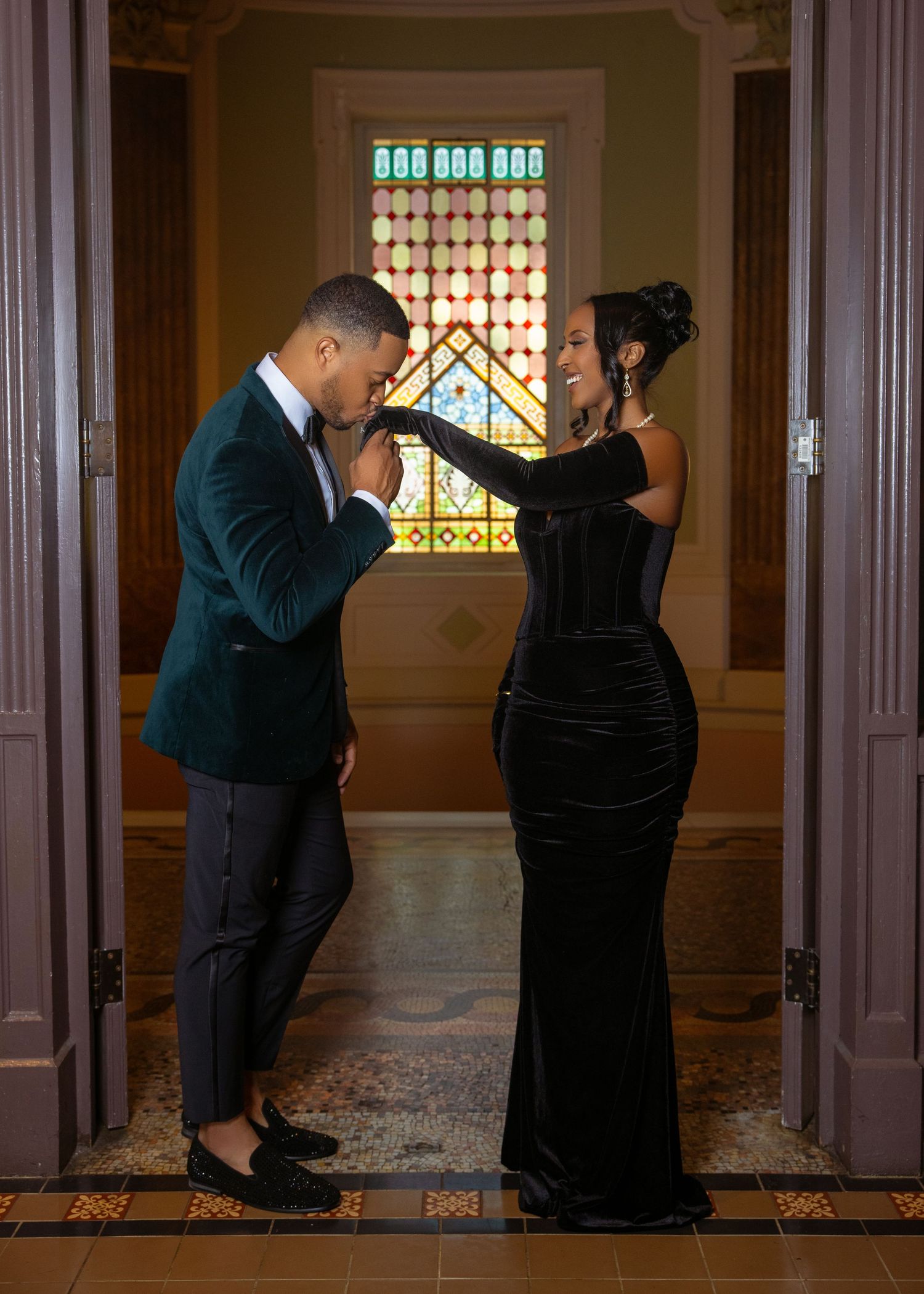 A couple in formal attire shares an intimate moment in a hallway with stained glass windows and wooden details.