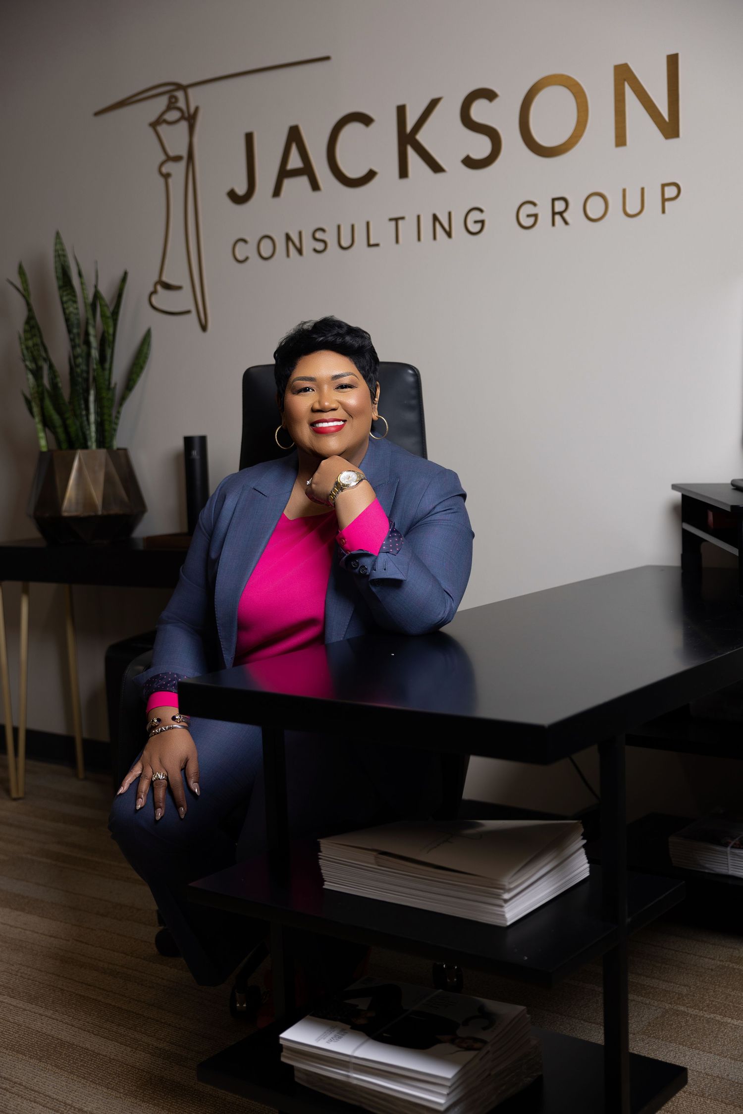Professional in a bright pink top and navy blazer sits at desk in modern office with Jackson Consulting Group signage.