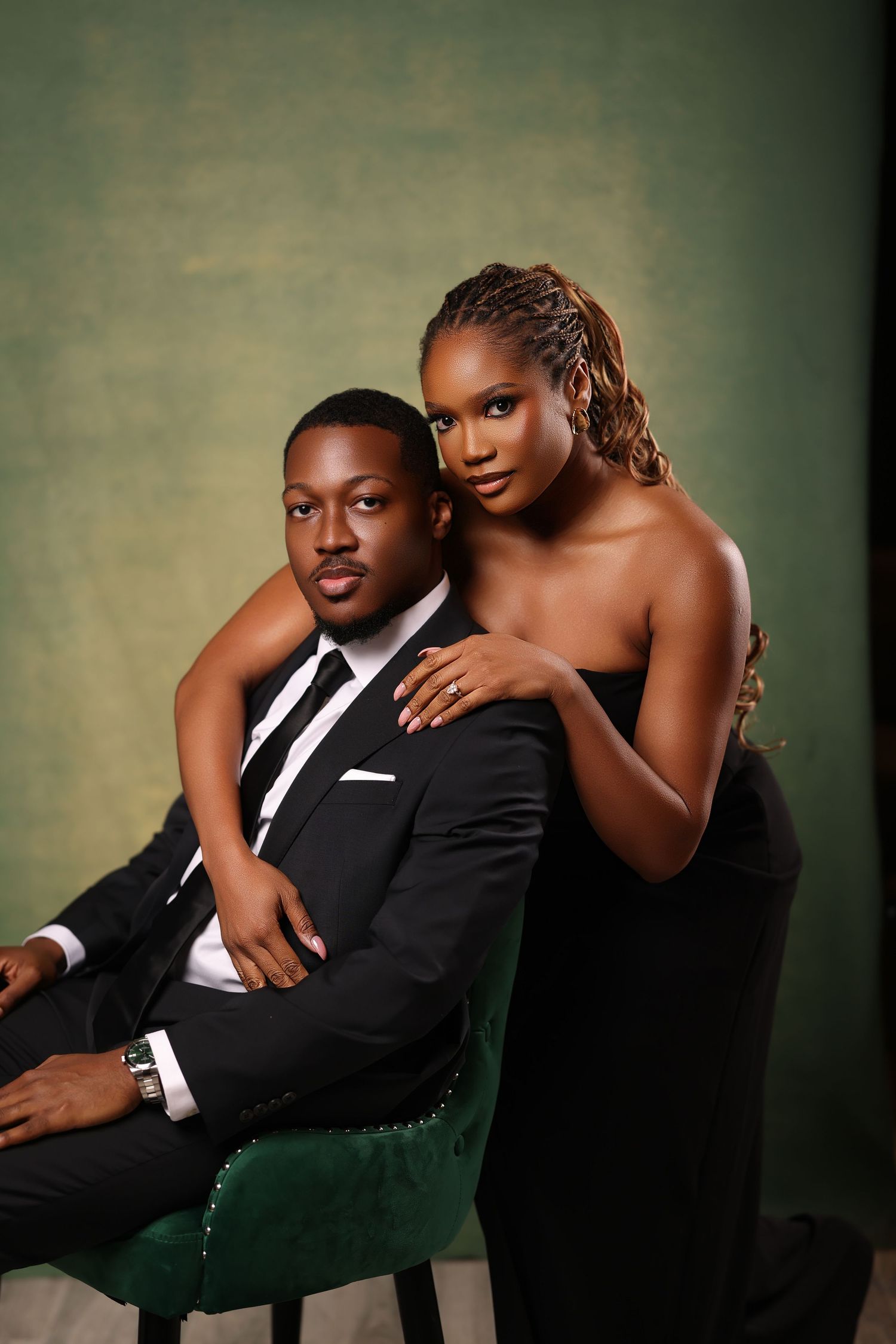 Elegant couple poses for formal portrait with person in black dress standing behind chair where partner sits in suit.