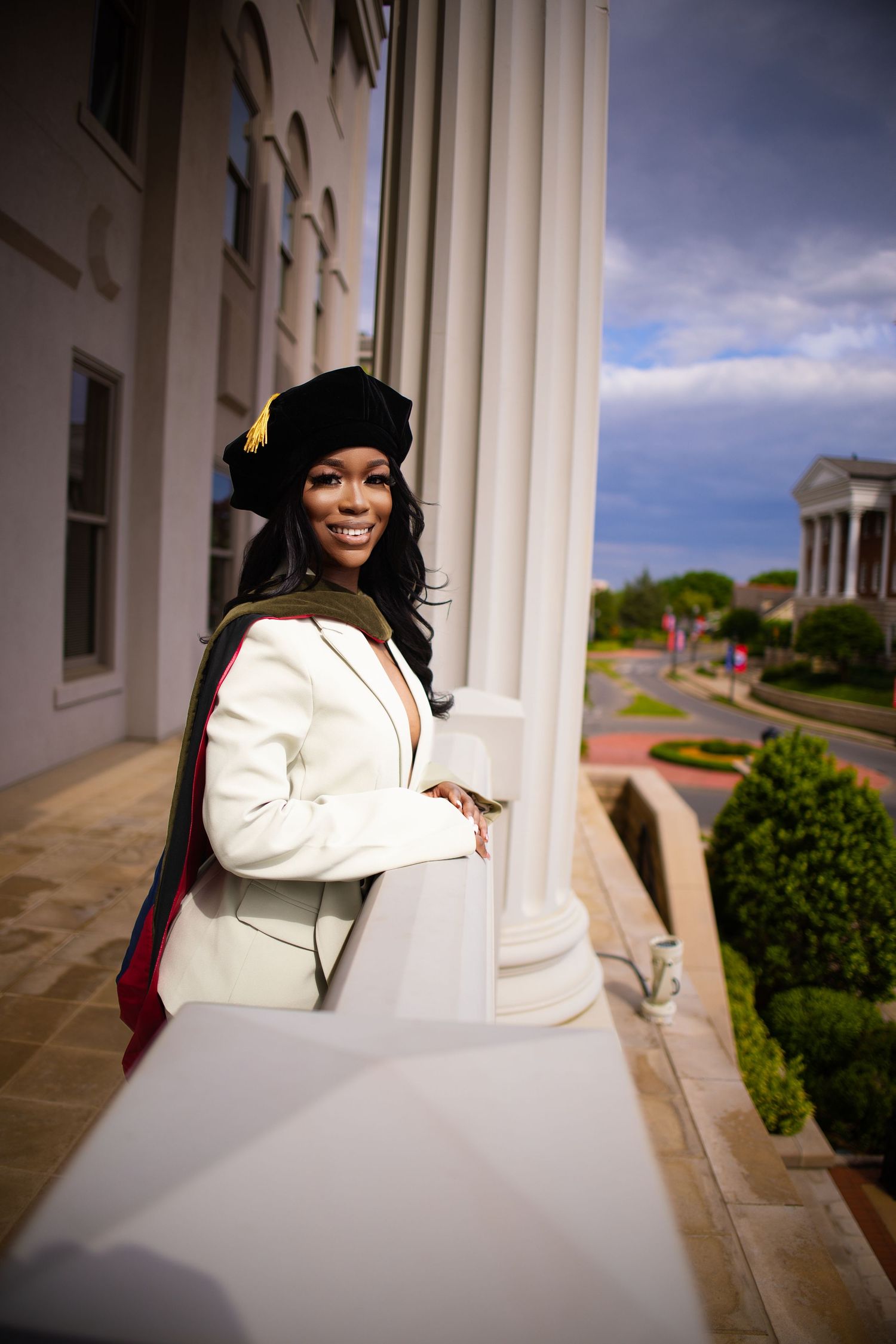 Graduate in academic regalia poses on a columned balcony of a stately university building under blue skies.