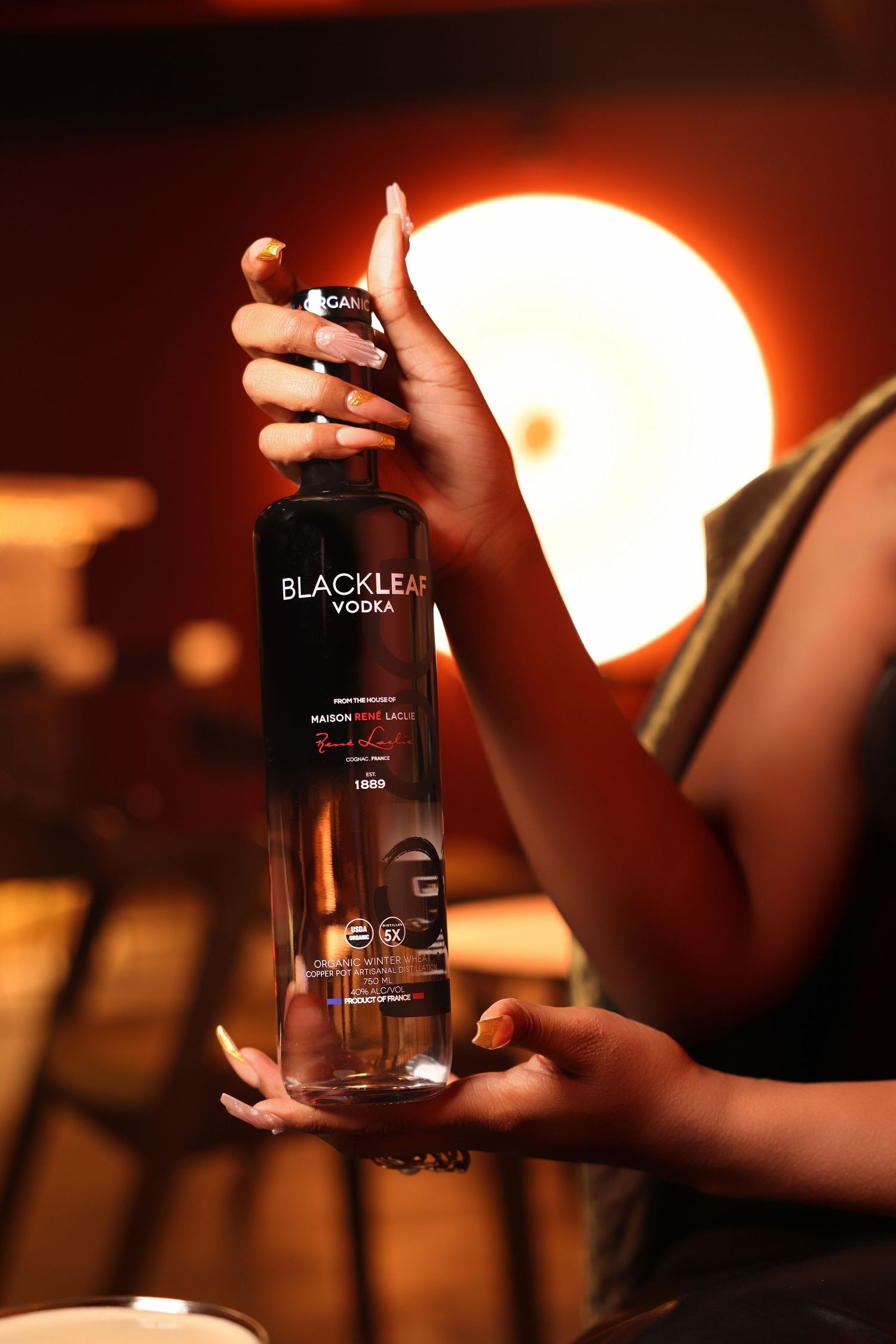 Hands holding Black Leaf vodka bottle in warm bar lighting.