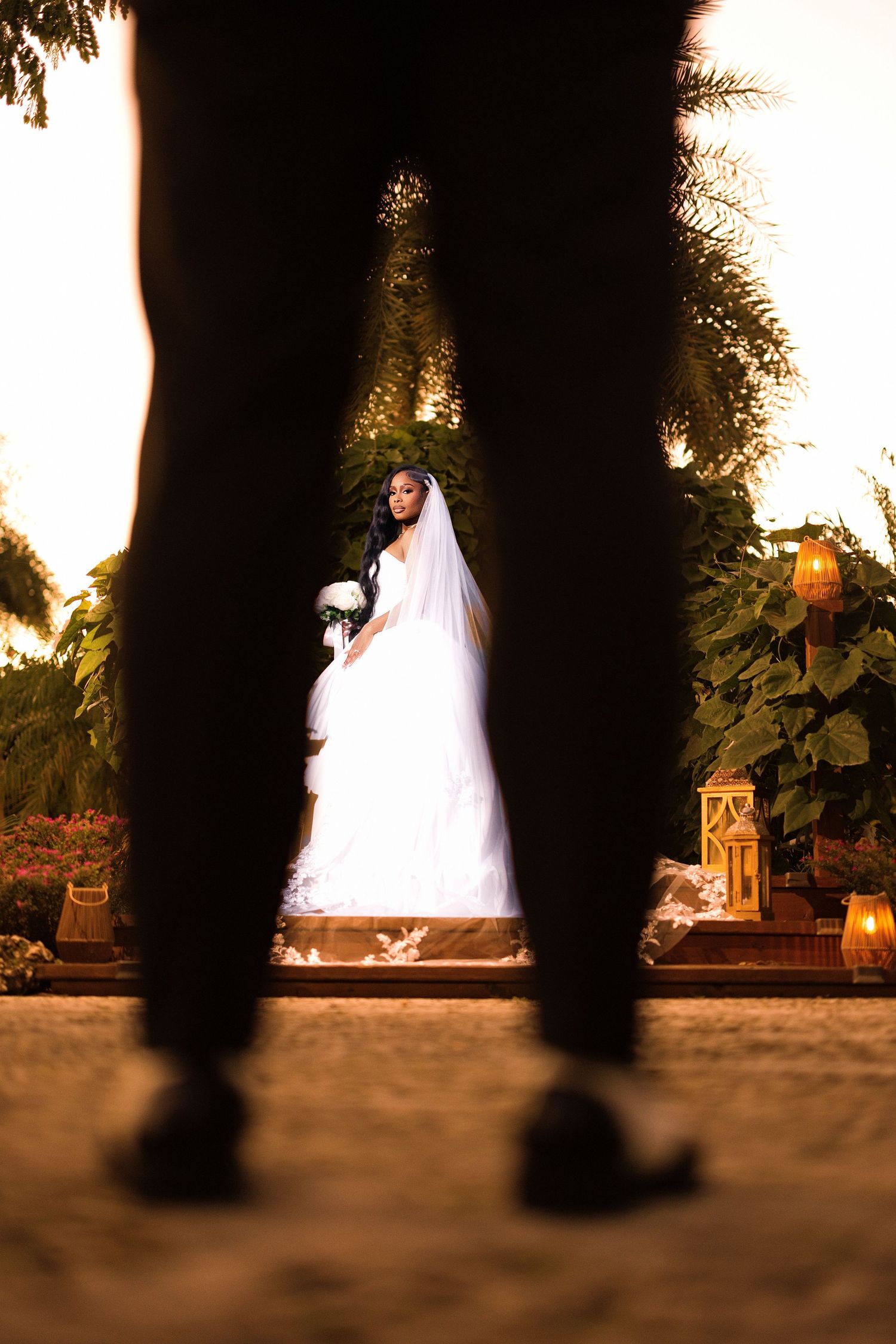 Silhouetted feet stand in the foreground while white wedding dresses glow softly in the background at sunset.