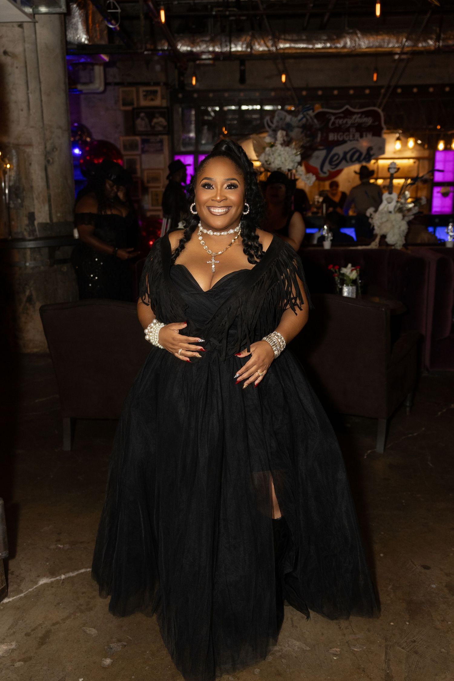 A person in a flowing black tulle gown poses at an indoor event venue with ambient lighting.