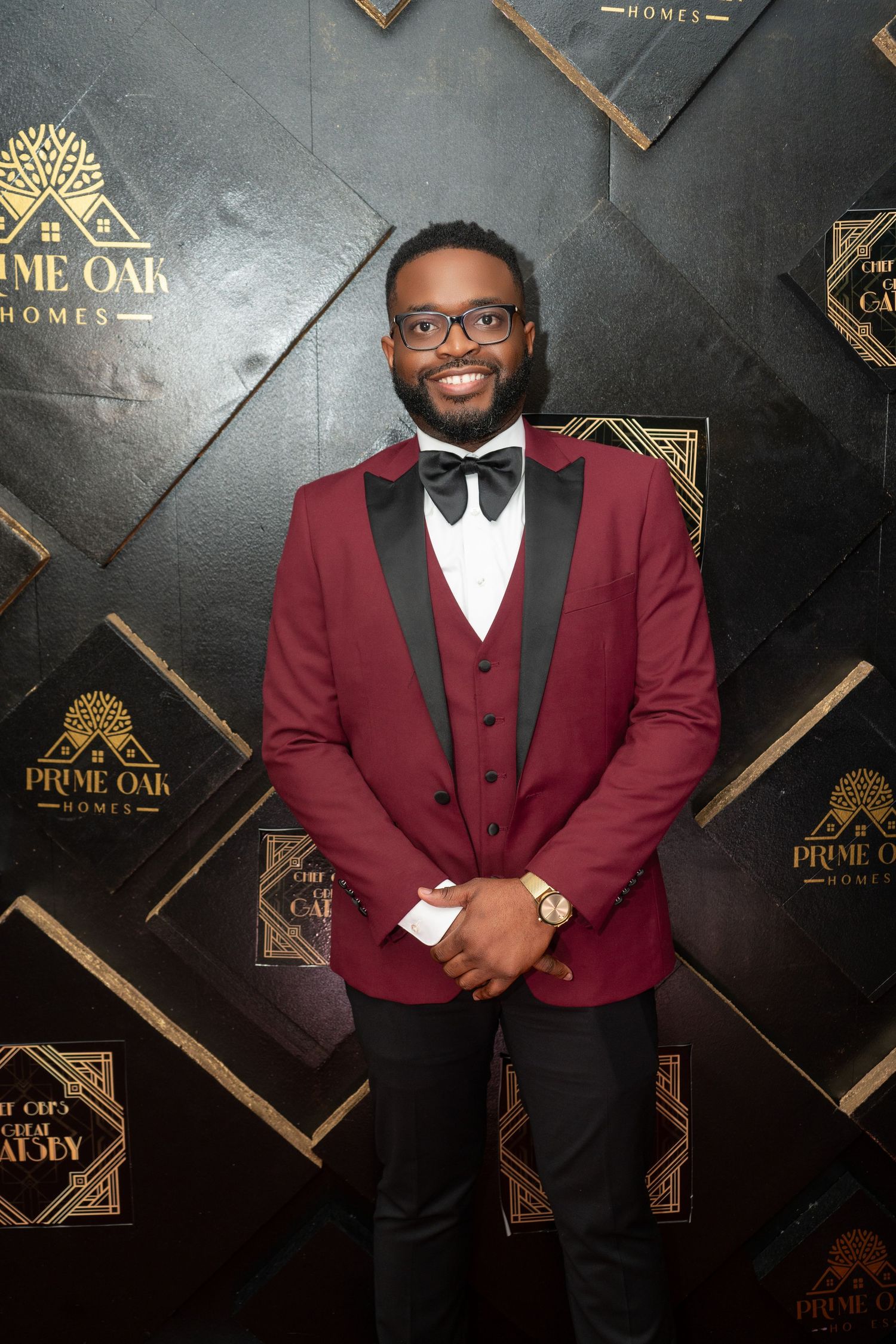A stylish burgundy three-piece tuxedo with black bow tie photographed against a geometric patterned backdrop.