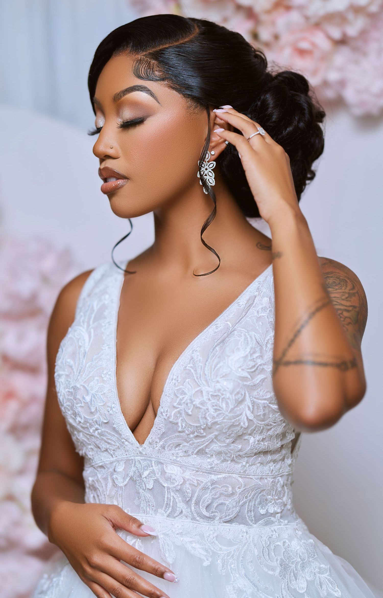 A bride in an elegant white lace wedding dress with intricate beading touches her sparkling earring against a floral backdrop.
