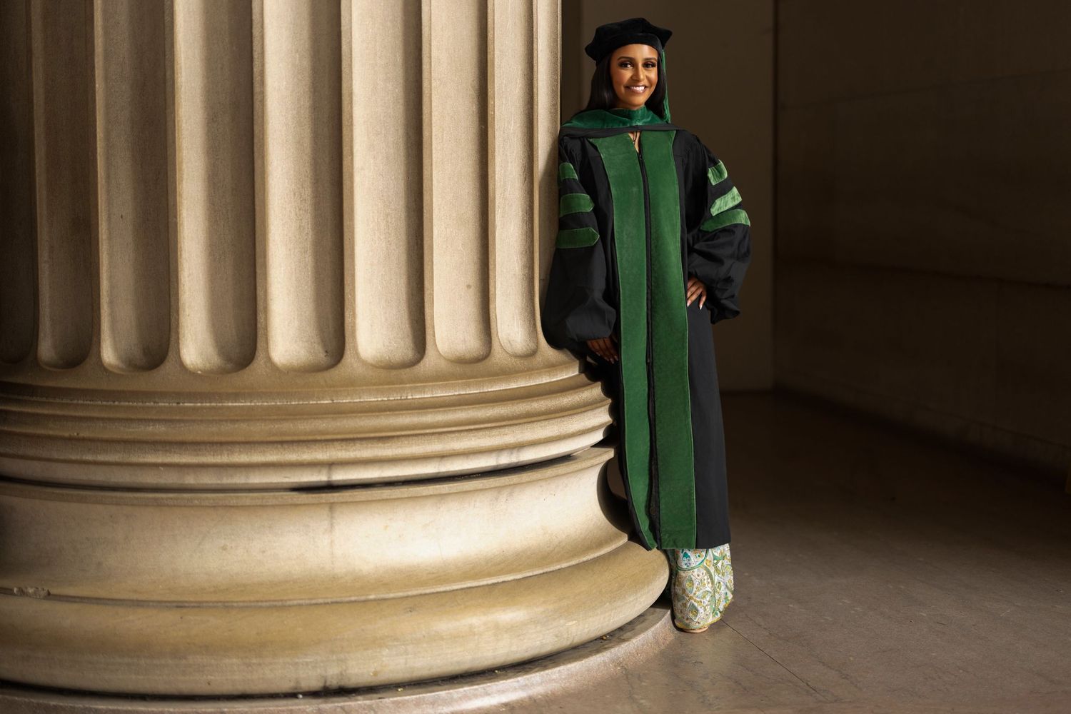 Person in green graduation gown stands between classical stone columns in dramatic lighting.
