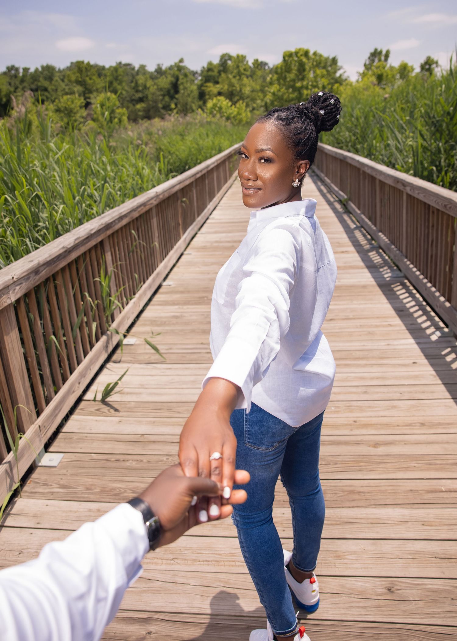 A couple holds hands on a wooden boardwalk surrounded by tall grass and trees on a sunny summer day.
