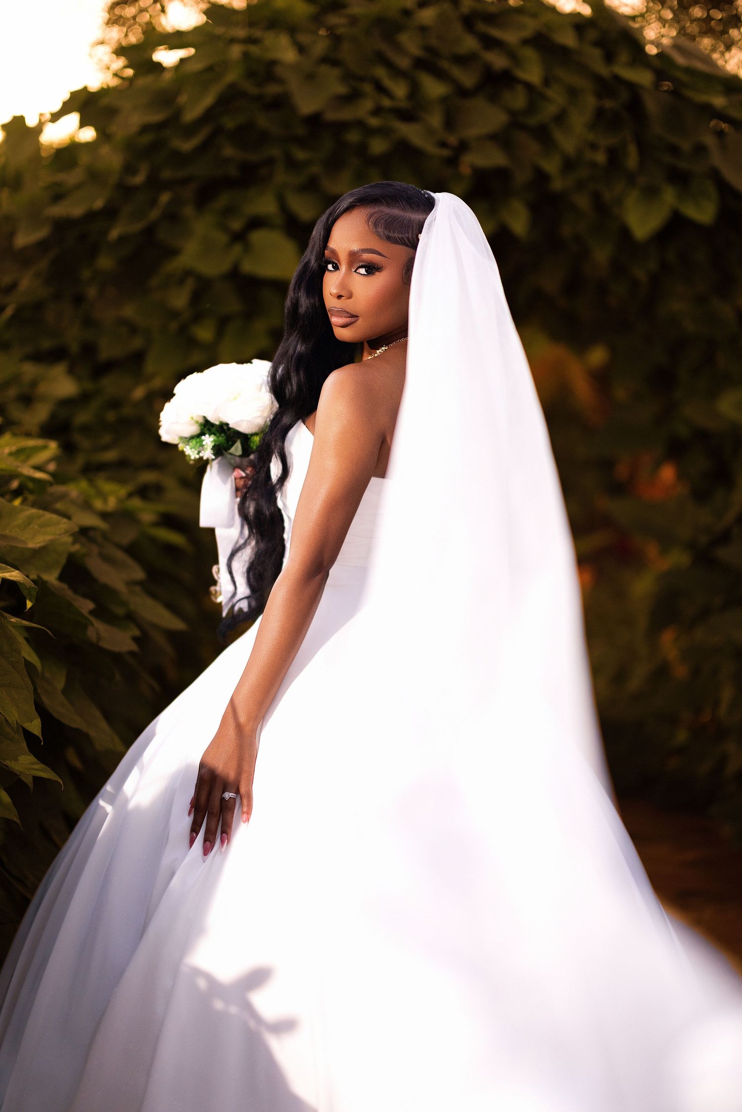 A bride in a flowing white wedding gown poses against a natural outdoor backdrop during golden hour.