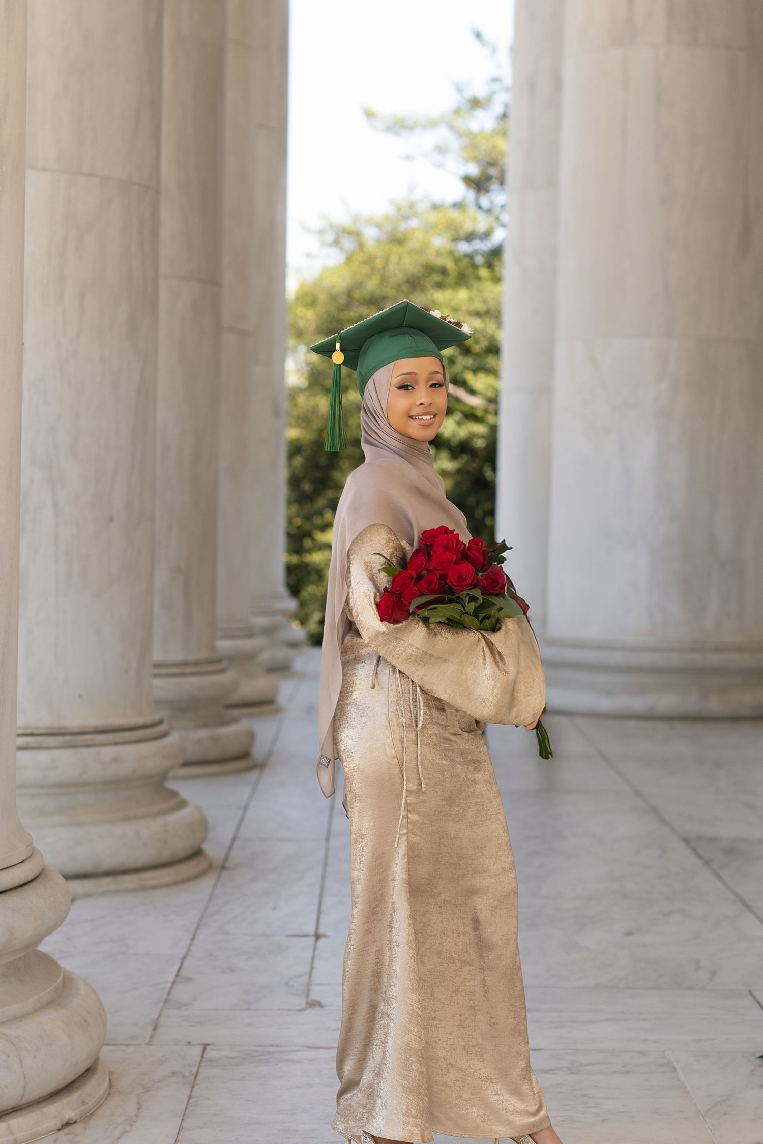 A person in a beige dress and hijab poses between classical stone columns with red roses.