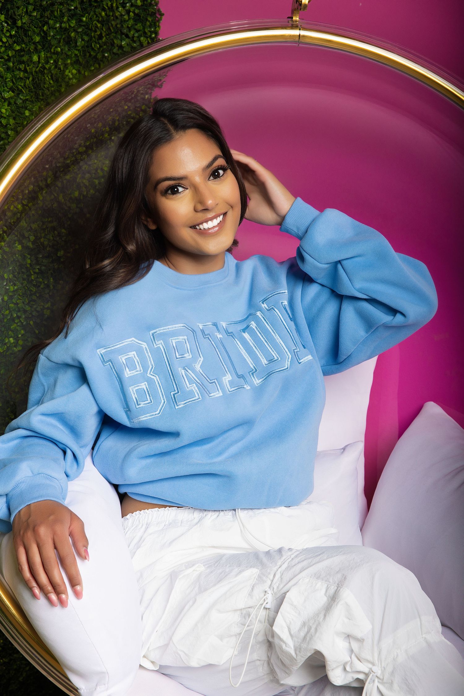 A person wearing a light blue BRNY sweatshirt and white pants poses in a circular chair against a pink and green backdrop.