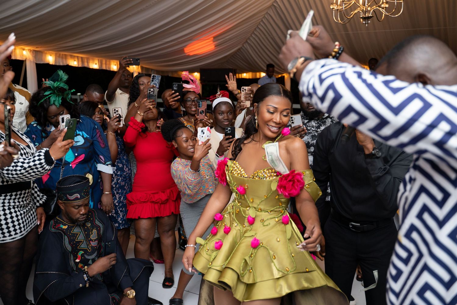 Energetic party scene with people dancing and celebrating in a decorated indoor venue with draped ceiling and chandelier.