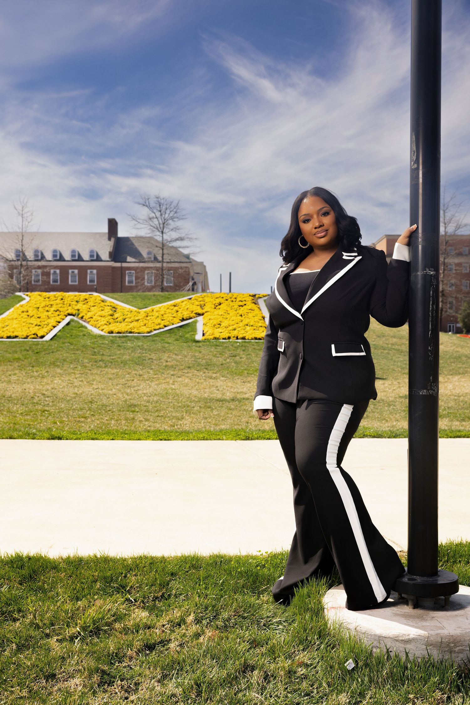 A person in a black and white outfit poses playfully in a grassy field on a sunny day against a bright blue sky.