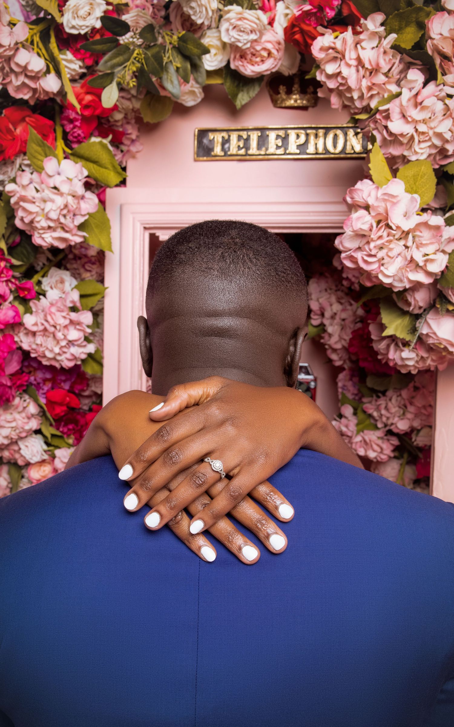 Hands with white manicure and ring cross behind back against pink telephone booth decorated with pink flowers.