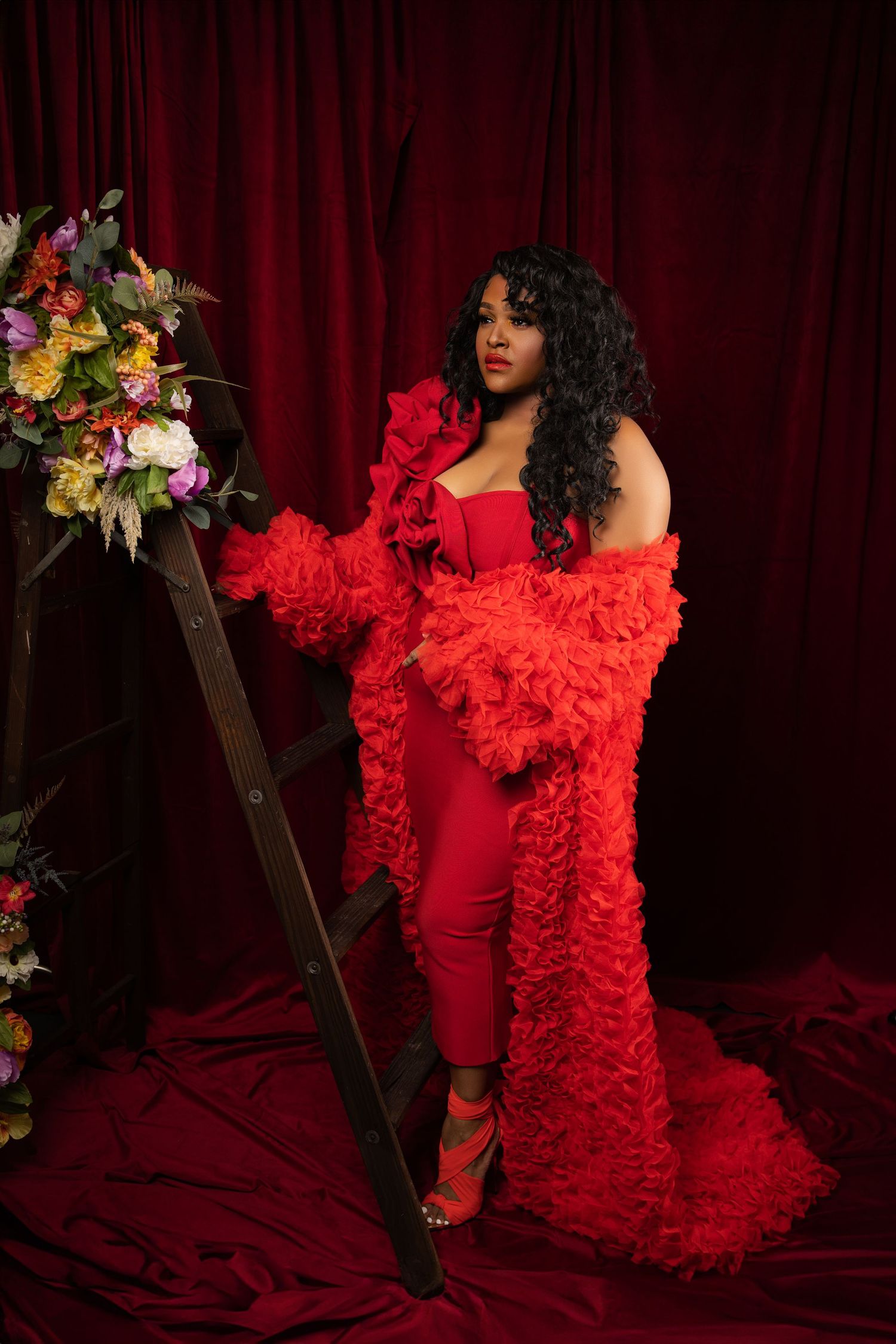 Glamorous fashion portrait featuring a red ruffled gown and floral arrangement against dark velvet curtains.