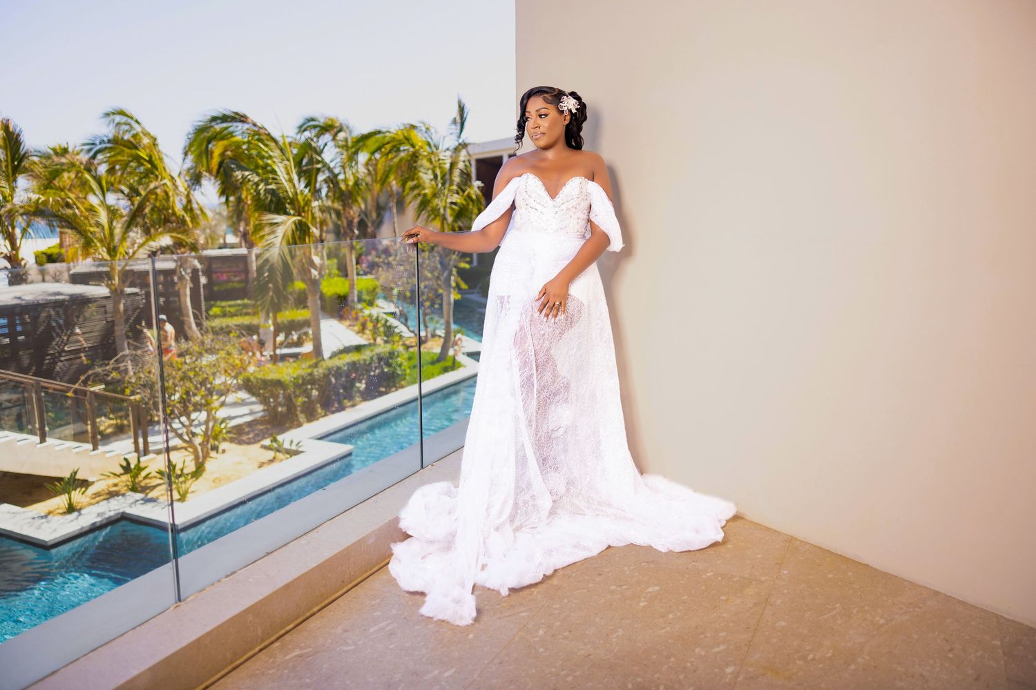 A bride in a flowing white wedding dress poses at a tropical resort with palm trees and ocean views in the background.