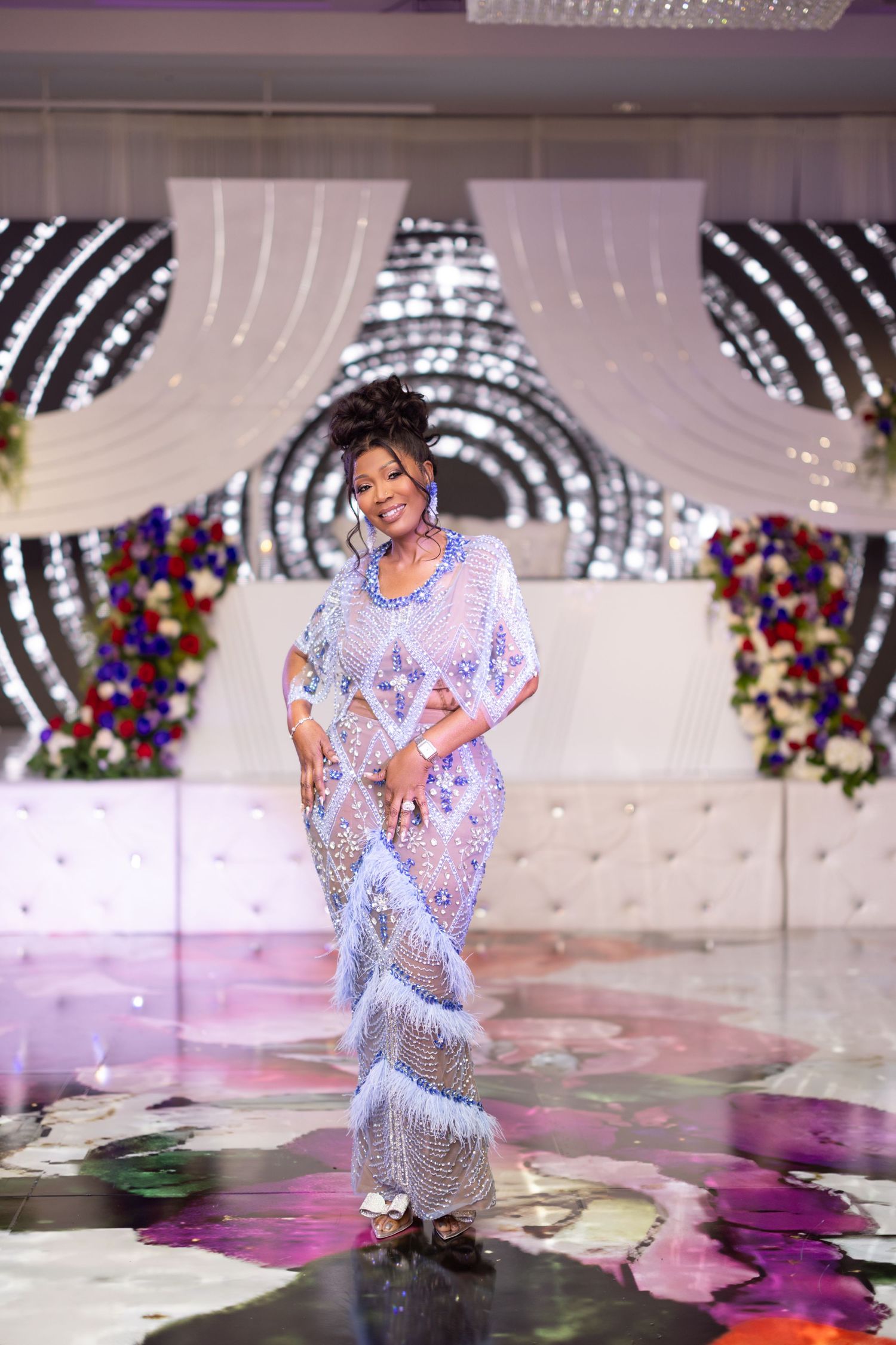 A performer in a shimmering white feathered dress dances on a reflective stage with colorful floral decorations.