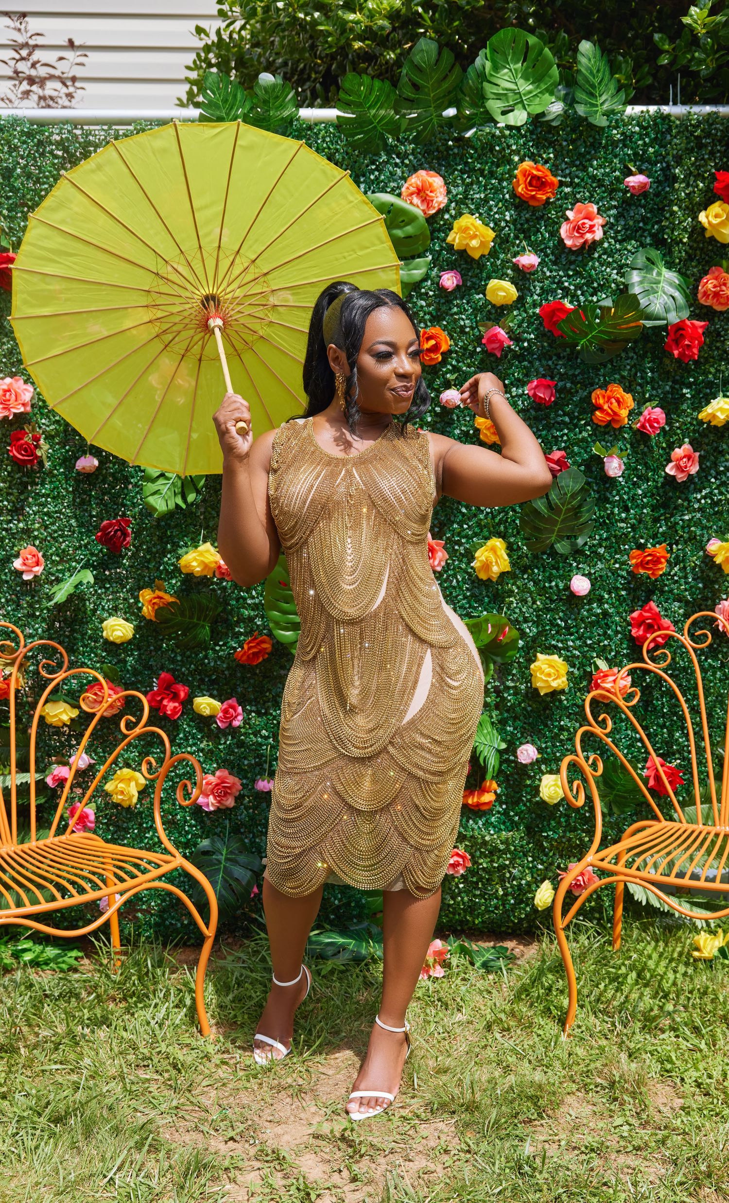 A gold sequined dress shimmers against a backdrop of colorful roses and orange garden furniture in an outdoor setting.
