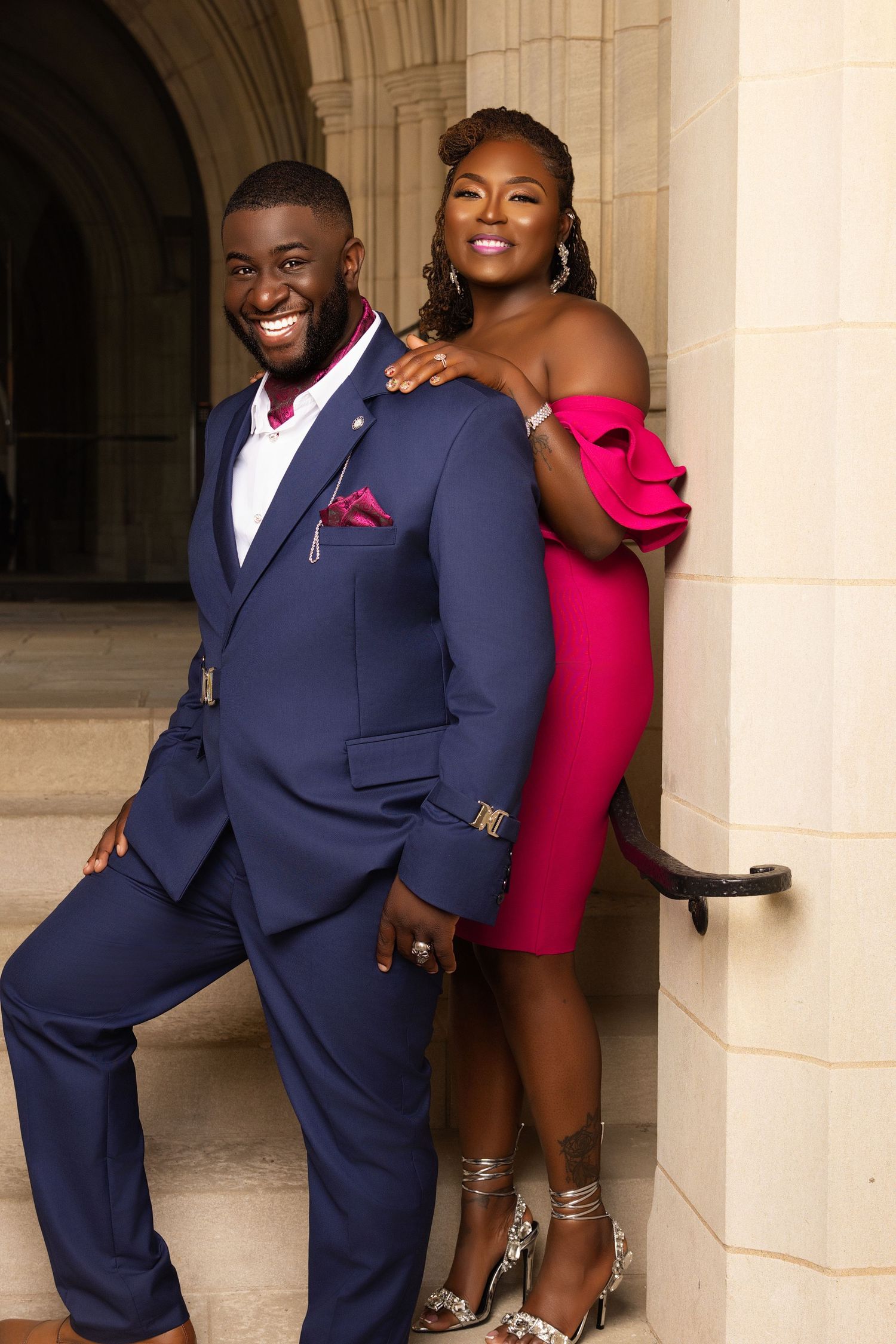 Couple in navy suit and pink dress poses playfully against stone columns.