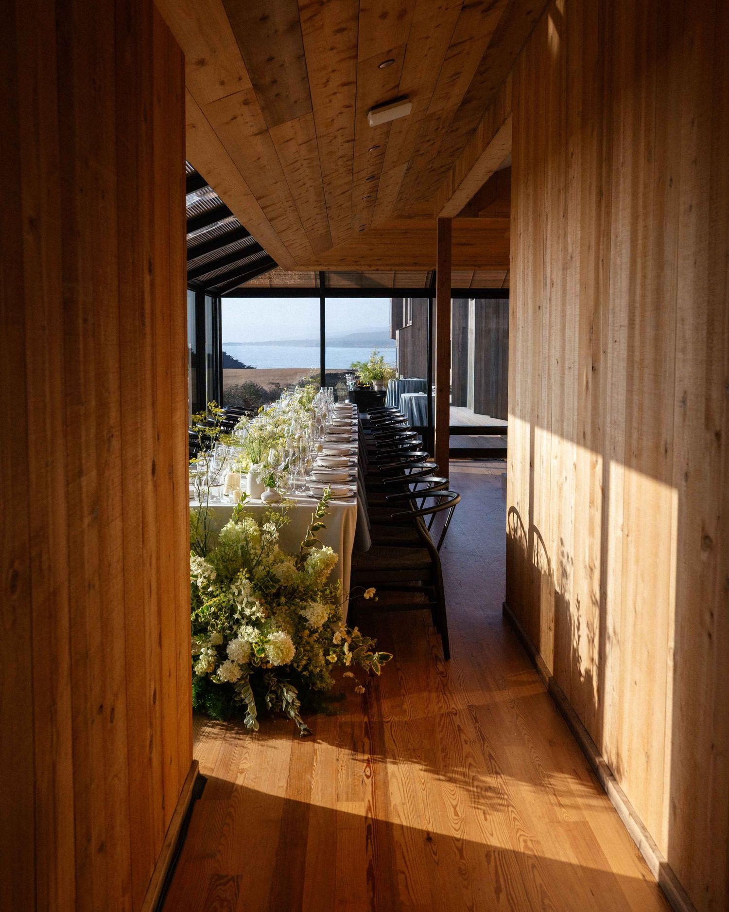 Elegant long dining table with white floral arrangements in a warm wood-paneled venue overlooking the ocean.