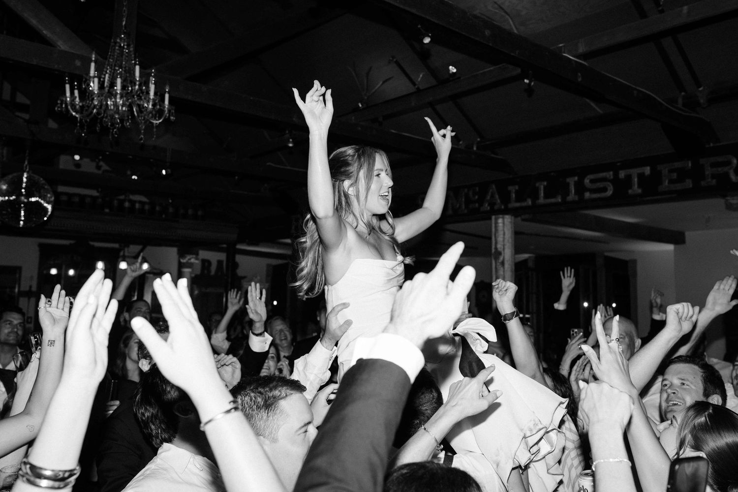 Bride crowd surfing at lively wedding reception, arms raised in celebration beneath a grand chandelier in black and white.