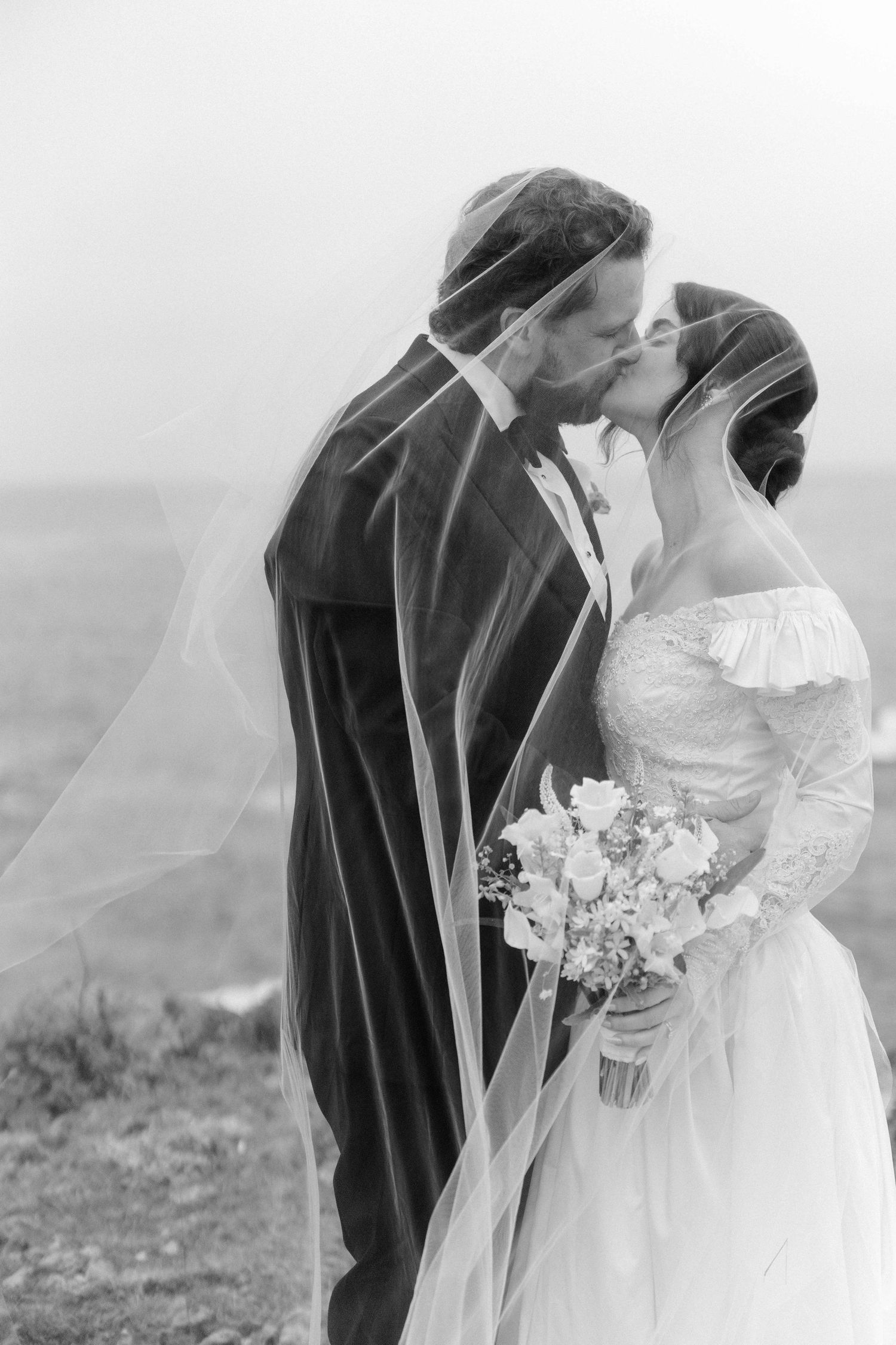 Black and white photo of a bride and groom kissing under a flowing veil outdoors, bride holding a bouquet.