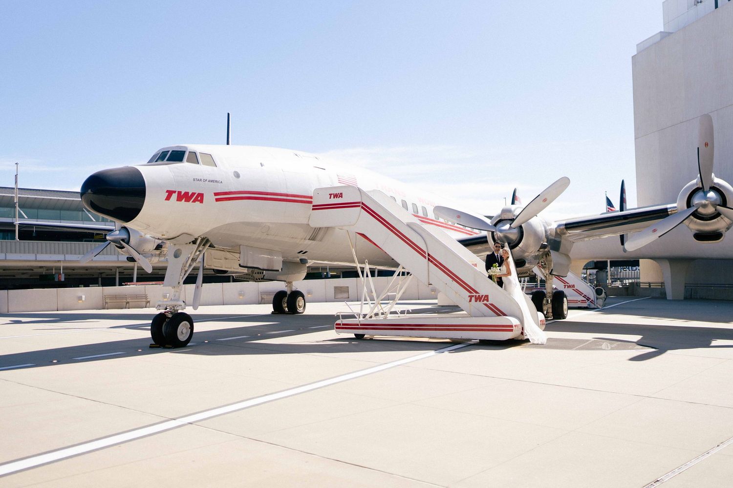 Vintage TWA propeller airplane on tarmac with boarding stairs, displayed outdoors under clear blue sky.