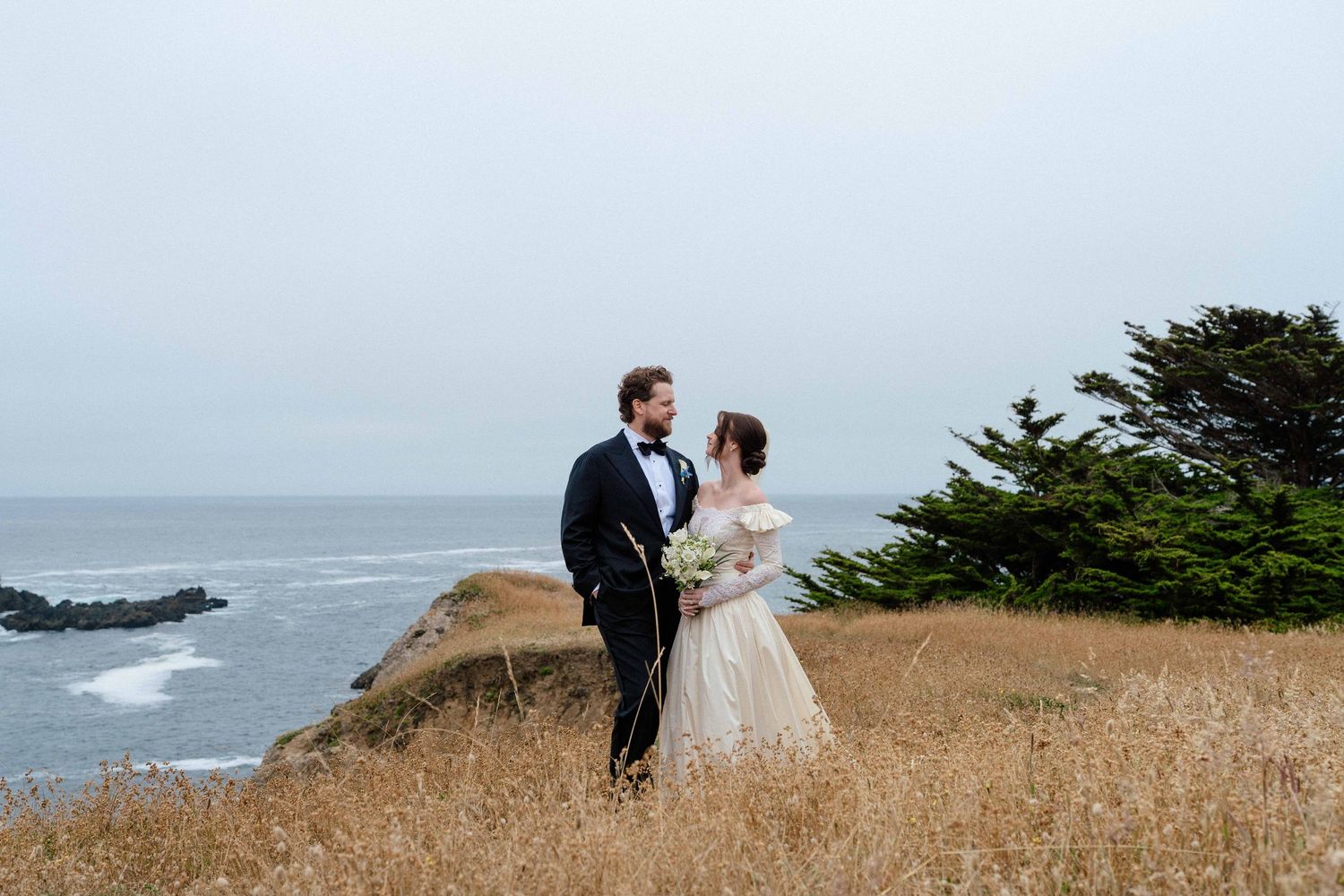 Couple in wedding attire standing in golden coastal grass with ocean views and evergreen trees on a misty day.