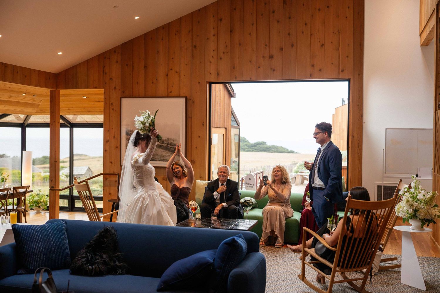 Bride holding bouquet aloft in a modern wooden venue with guests seated on blue sofas during a wedding celebration.