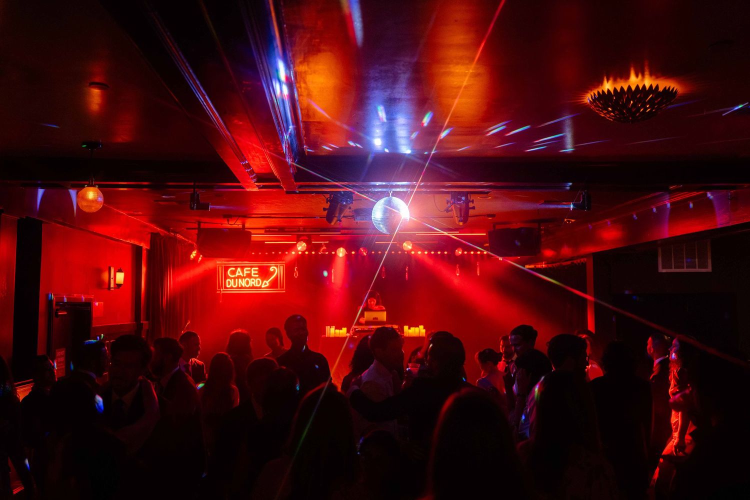 Crowded nightclub with red neon lights, laser beams, and a disco ball illuminating dancers on the floor.