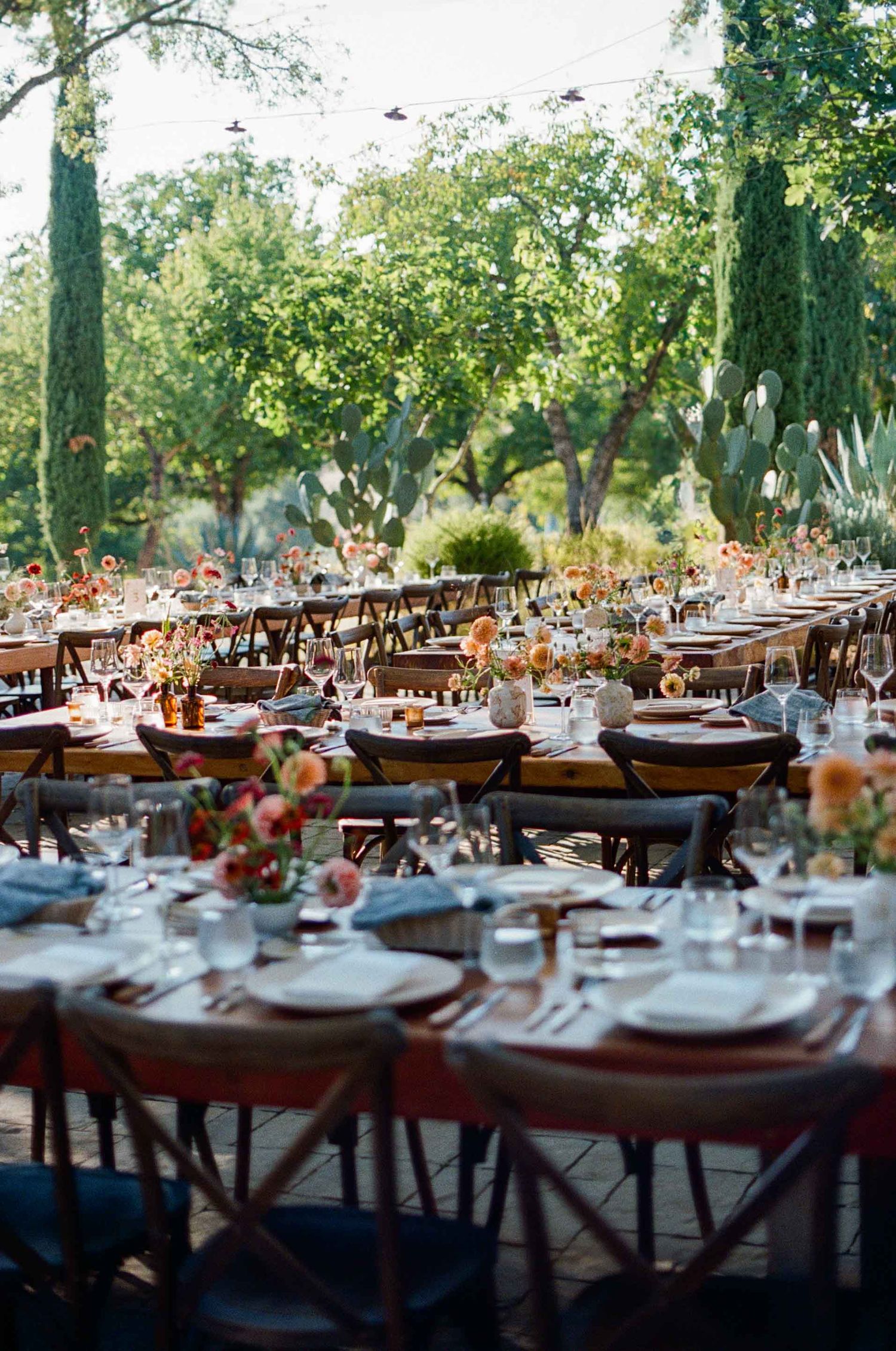 Elegant outdoor wedding reception with long wooden tables, floral centerpieces, and lush greenery.