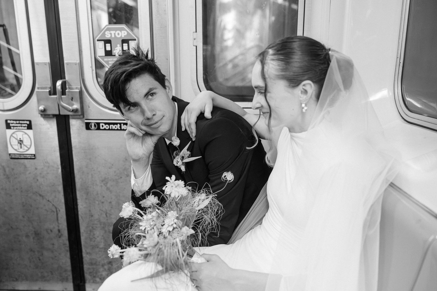 Black and white photo of a bride and groom riding a subway train, bride holding wildflowers bouquet.