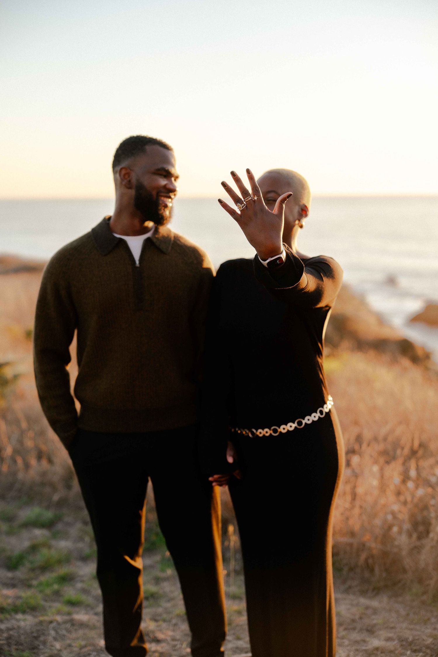 Couple standing outdoors at sunset near the ocean, woman showing engagement ring on her hand.