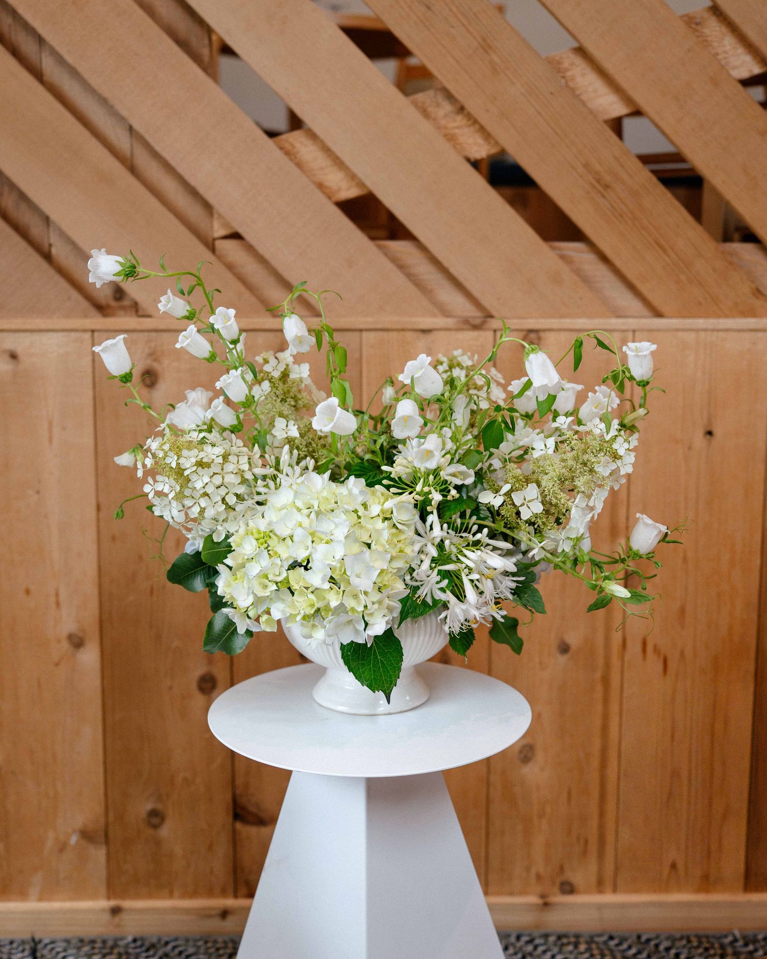 White hydrangeas and bell flowers arranged in a white vase on a pedestal table against a wooden wall.
