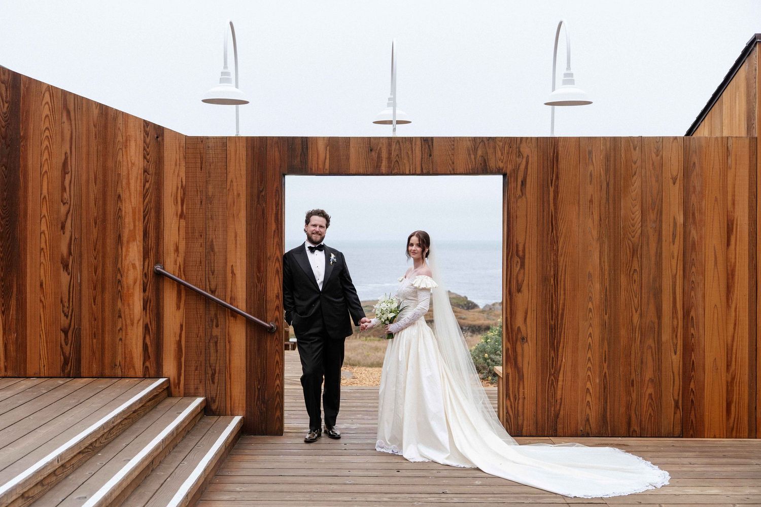 Bride in white off-shoulder gown and groom in black tuxedo stand in wooden arch with ocean views at outdoor wedding venue.