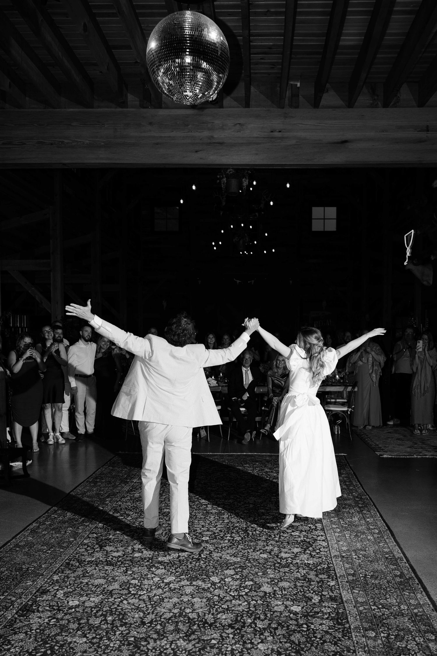Black and white photo of two brides holding hands with arms raised on dance floor under disco ball at wedding reception.