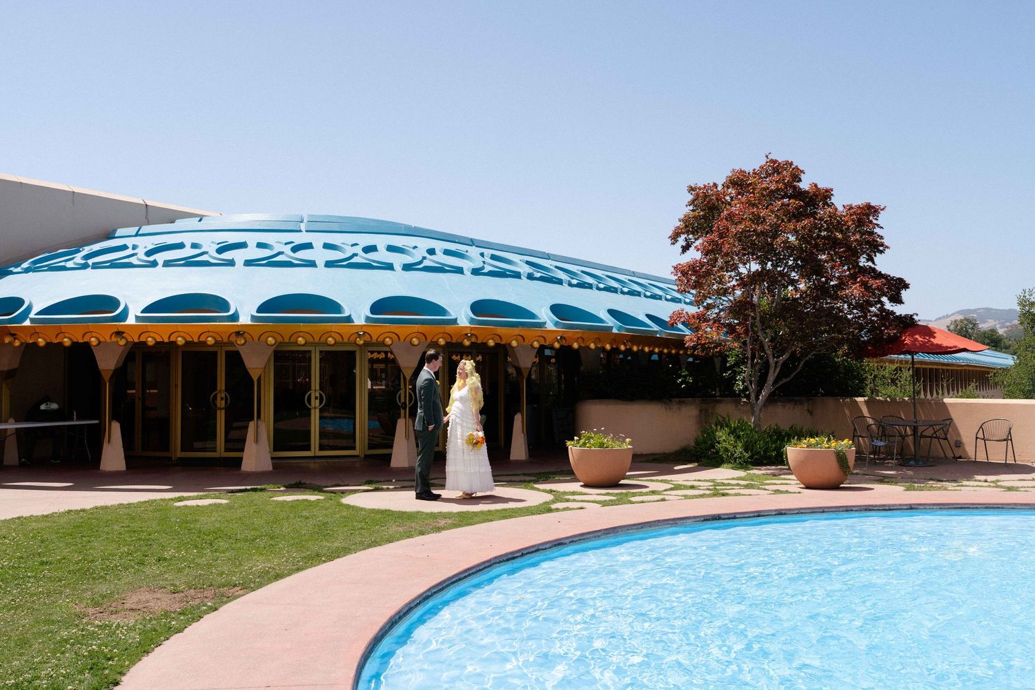 Bride and groom stand poolside near a colorful domed venue building with blue trim on a sunny day.