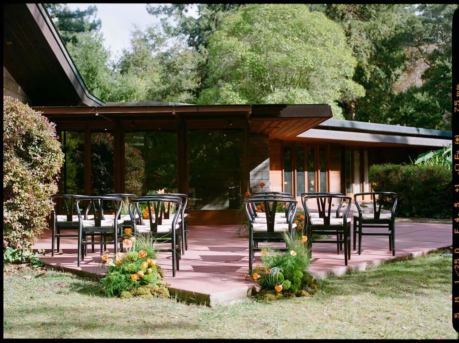 Outdoor mid-century modern wedding ceremony setup with black wishbone chairs on a brick patio surrounded by lush greenery.