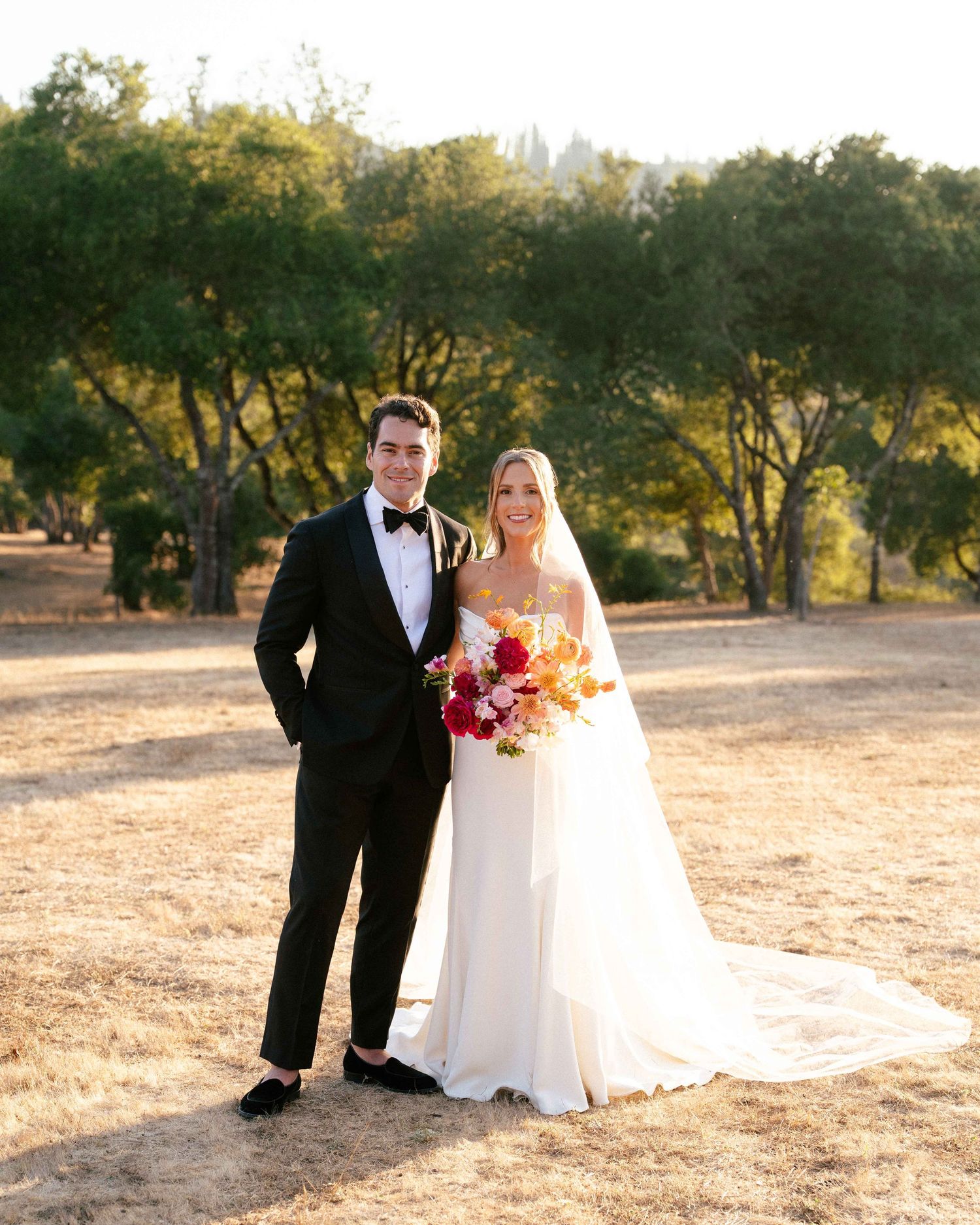 Bride and groom posing together outdoors, she holds a vibrant bouquet of orange and red flowers.
