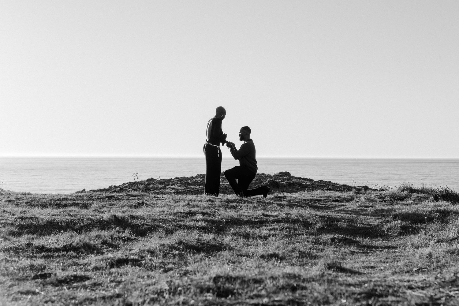 Black and white photo of a couple during a romantic marriage proposal on a grassy cliff overlooking the ocean.