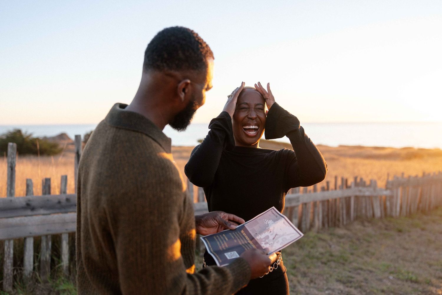 Couple sharing a joyful moment at sunset on a beach, woman laughing with hands on head while man holds a tablet.