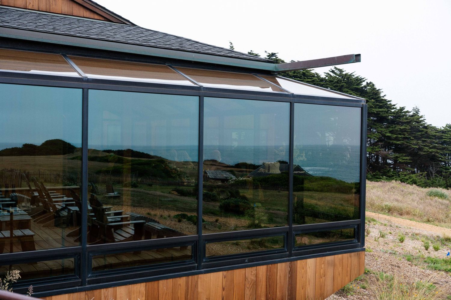 Modern glass and cedar building with panoramic ocean views reflecting green coastal landscape under overcast skies.