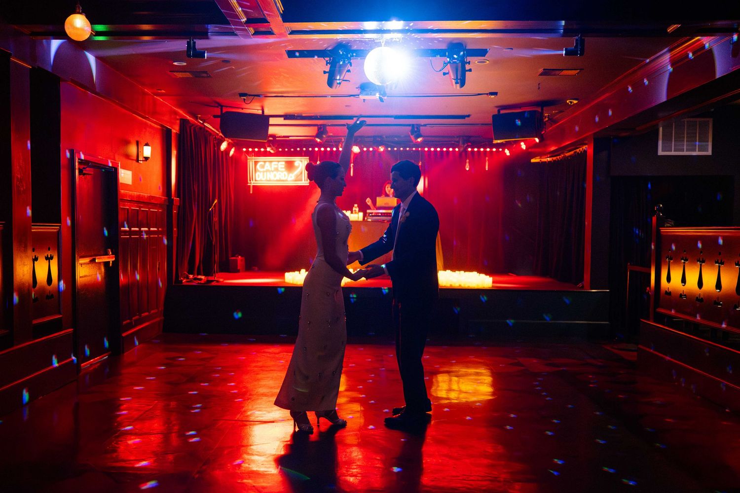 Couple dancing romantically in a dimly lit red-hued venue with stage lighting overhead.