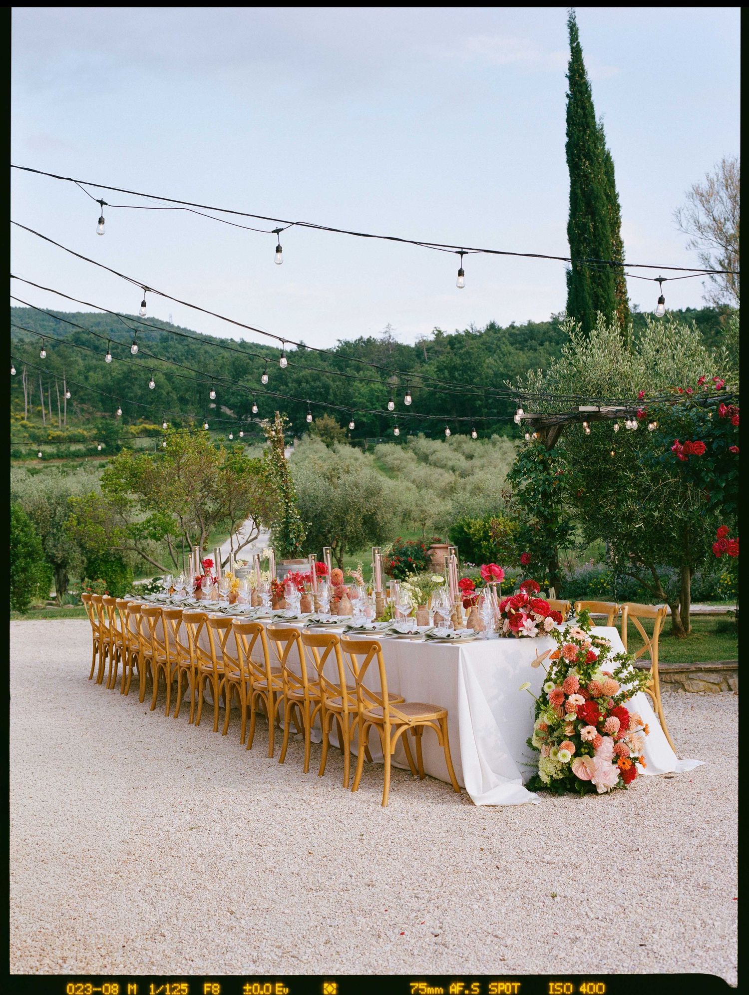 Elegant outdoor wedding reception table with floral arrangements, string lights, and cypress trees in Tuscany.