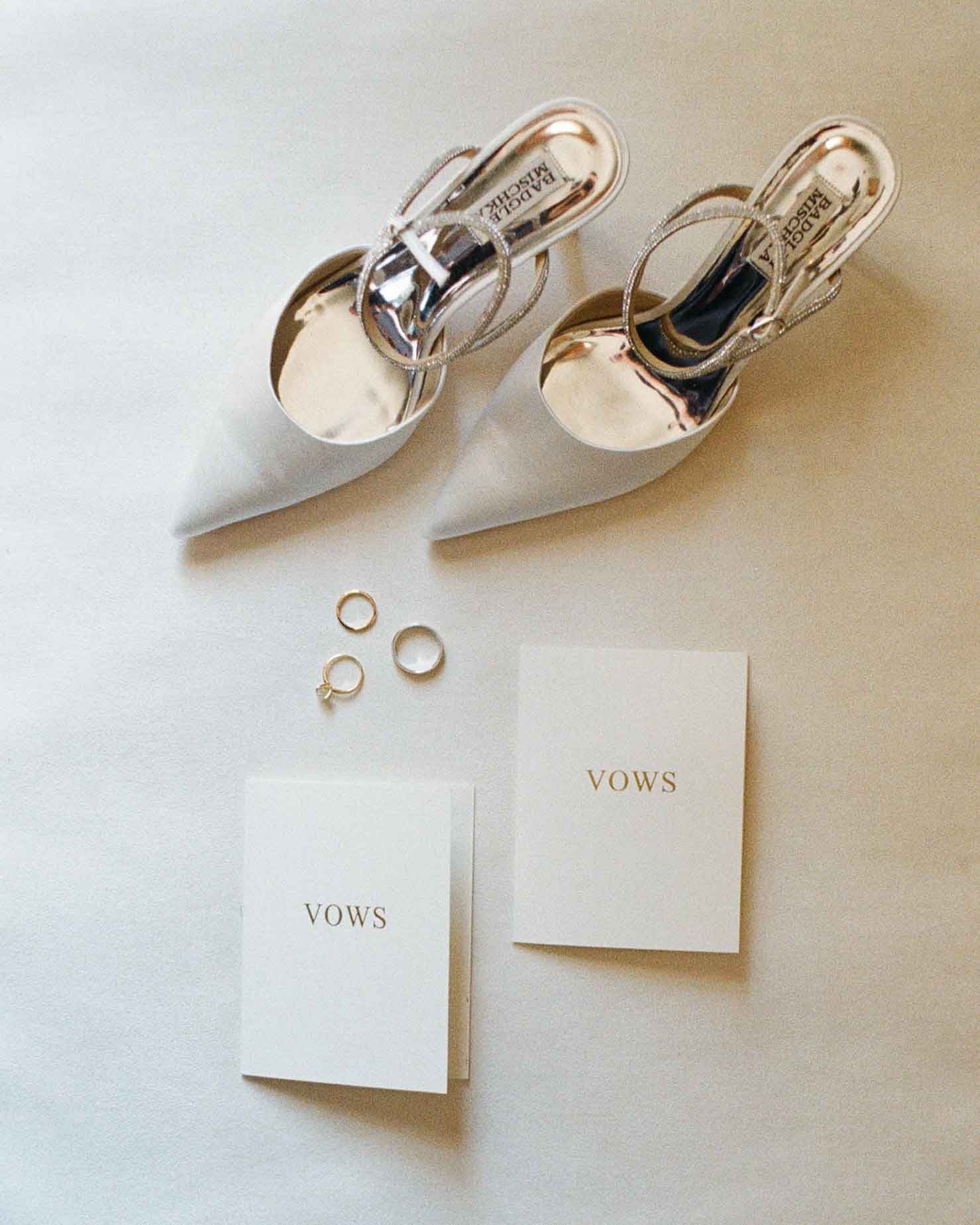 White bridal heels, gold rings, and two vow booklets arranged on a soft white surface.