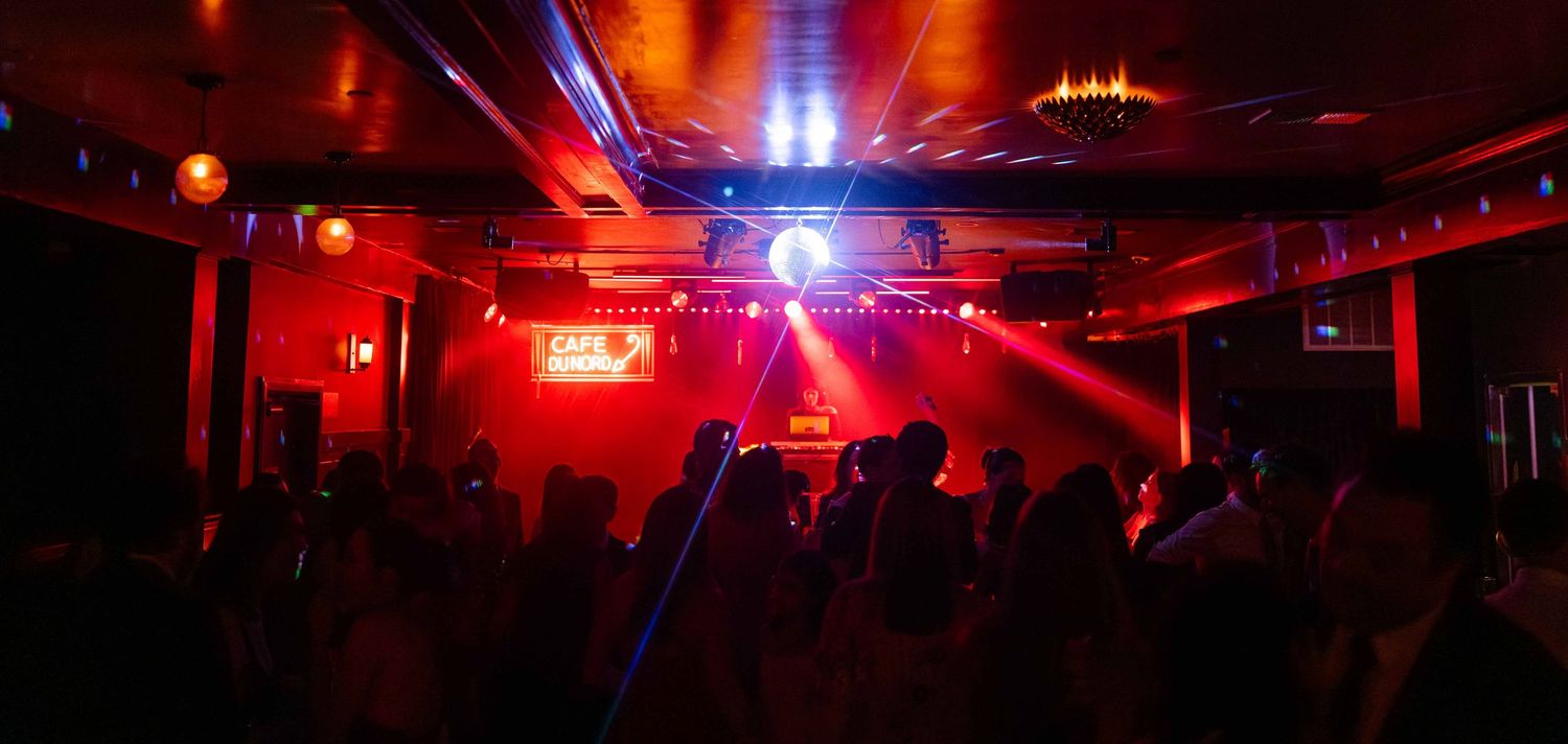 Crowded nightclub dance floor illuminated by red lights and a disco ball with colorful laser beams.