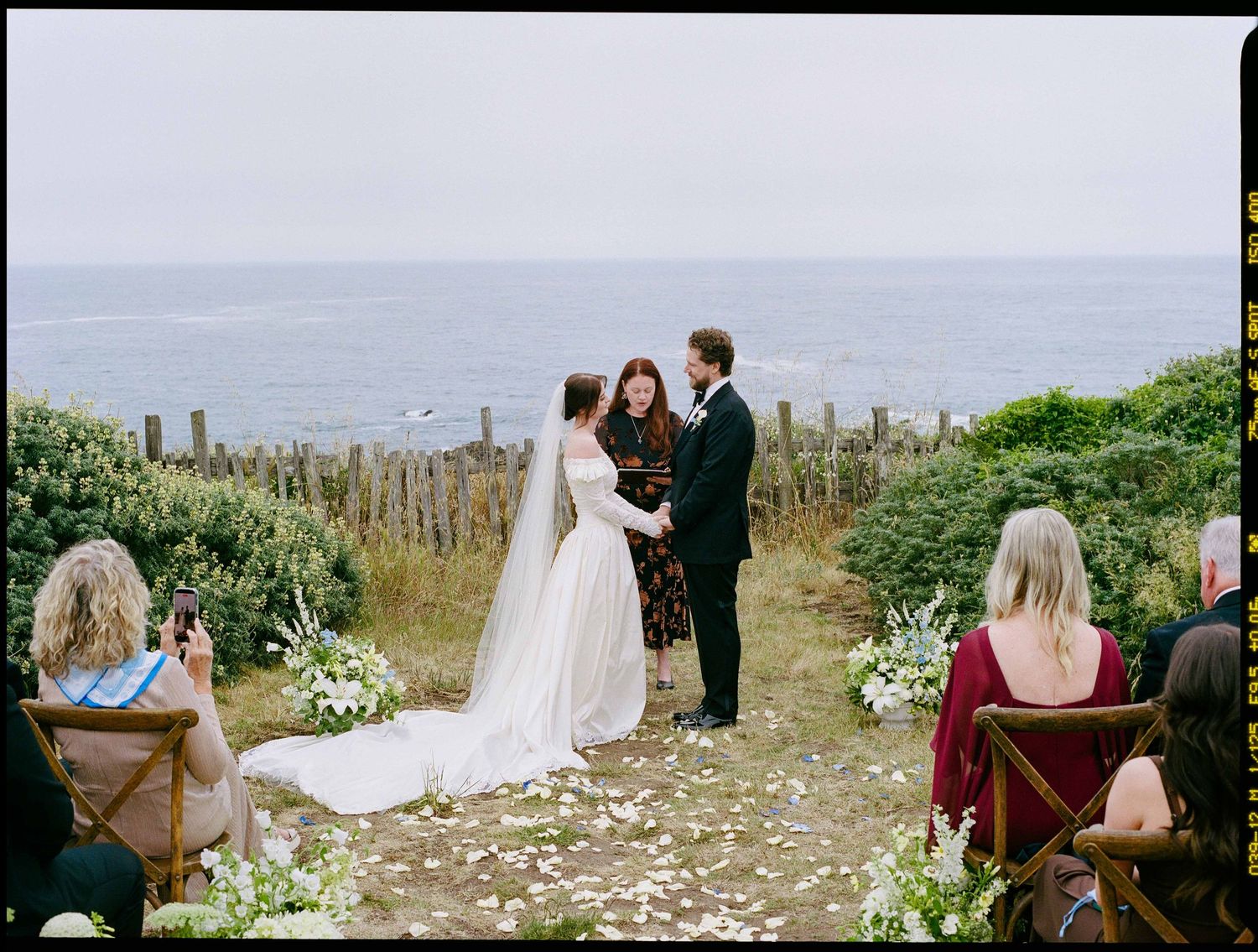 Outdoor coastal wedding ceremony with bride and groom exchanging vows by the ocean, surrounded by white floral arrangements.